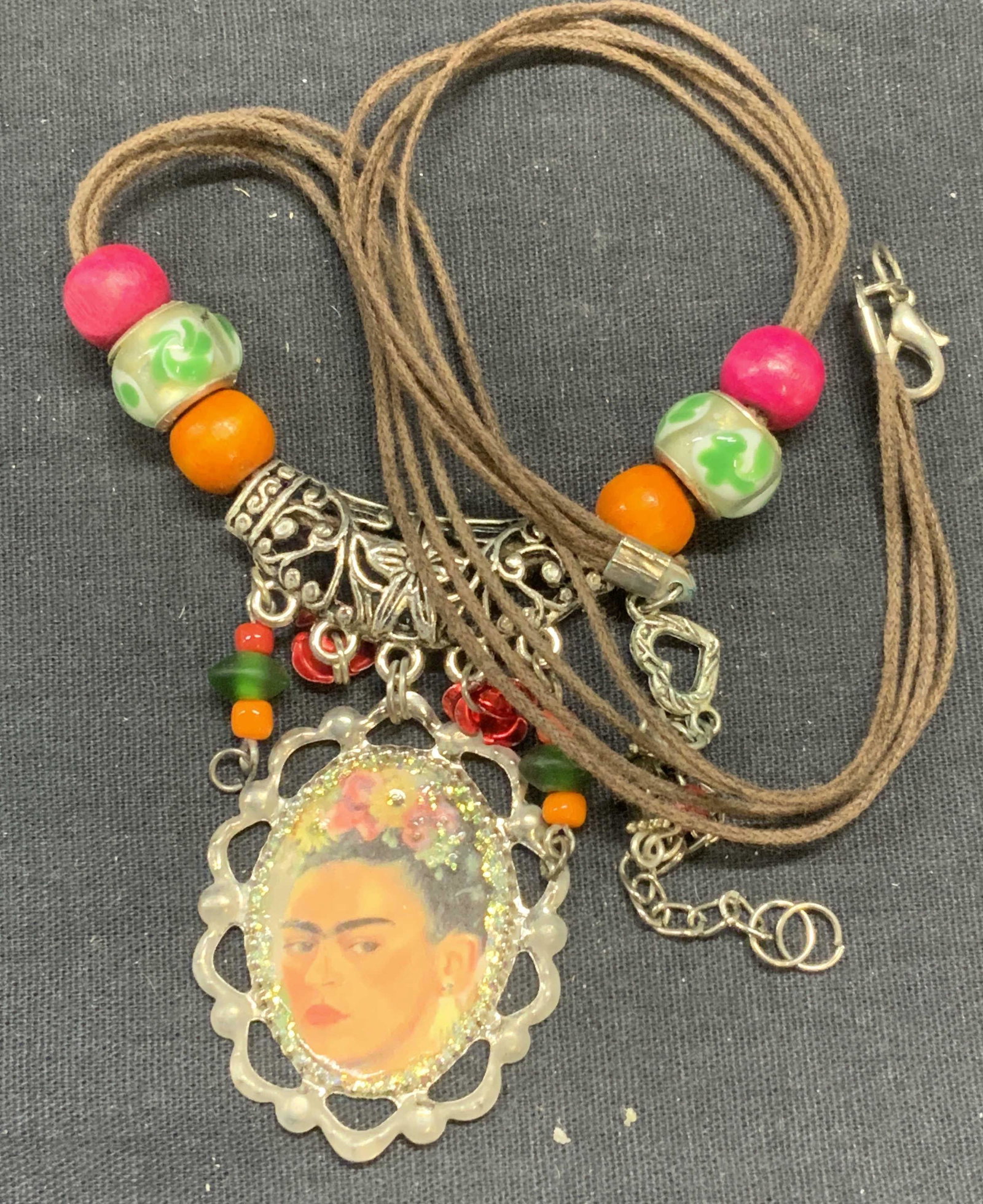 Beaded Frida Kahlo Pendant Necklace: Beaded Frida Kahlo pendant necklace on brown cord. Includes brown jewelry pouch. Measures approx. 19.5 inches long, pendant approx. 1 5/8 x 1 3/8 inches. Some surface wear consistent with exposure. Es
