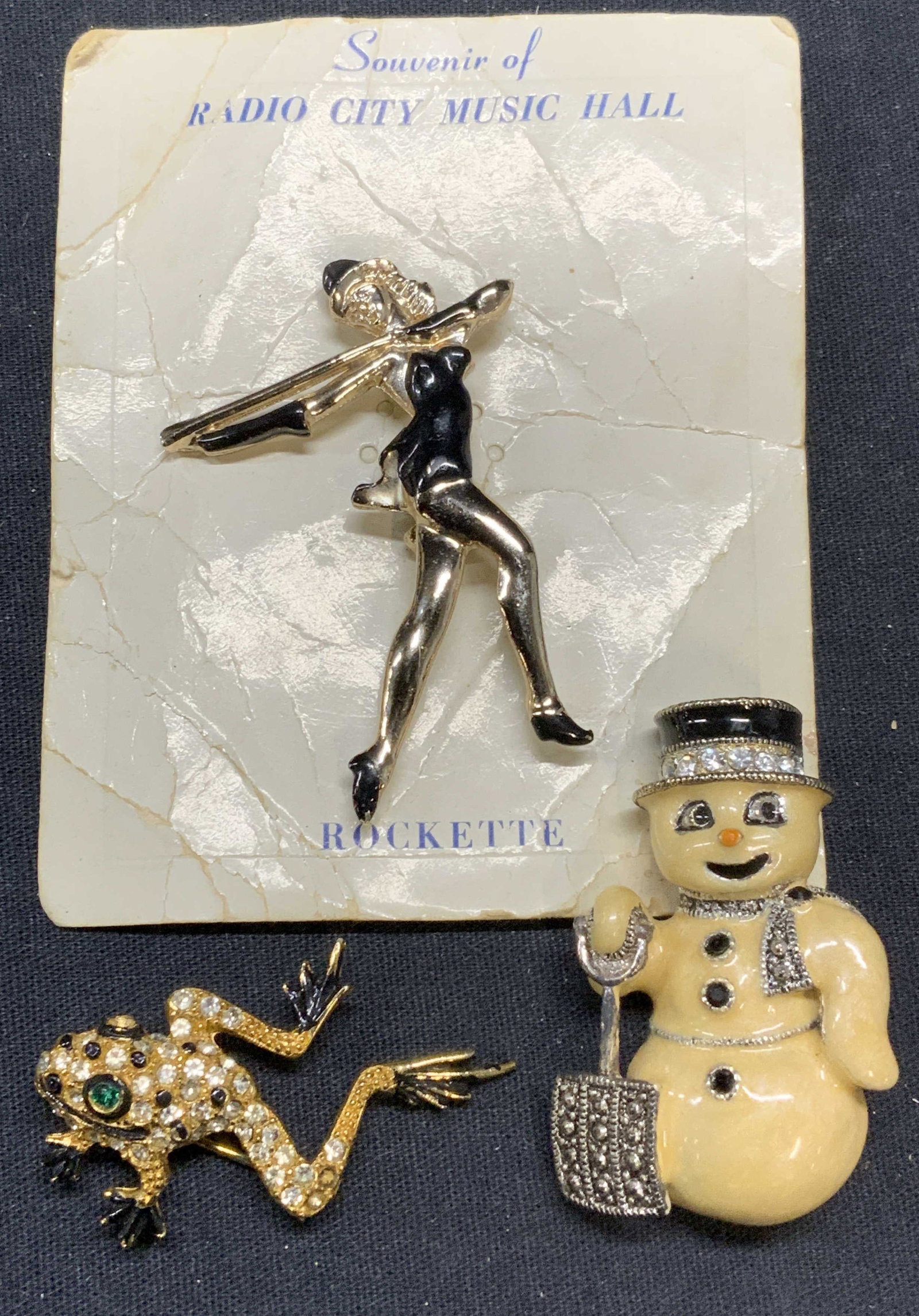 Radio City Rockette, Eisenberg, Frog, 3 Brooches: Lot of 3 jewelry pieces. Radio City Music Hall souvenir Rockette enameled brooch. Measures approx. 2.25 x 1.5 inches. Eisenberg enameled snowman brooch. Measures approx. 1 7/8 x 1 1/8 inches. Designer