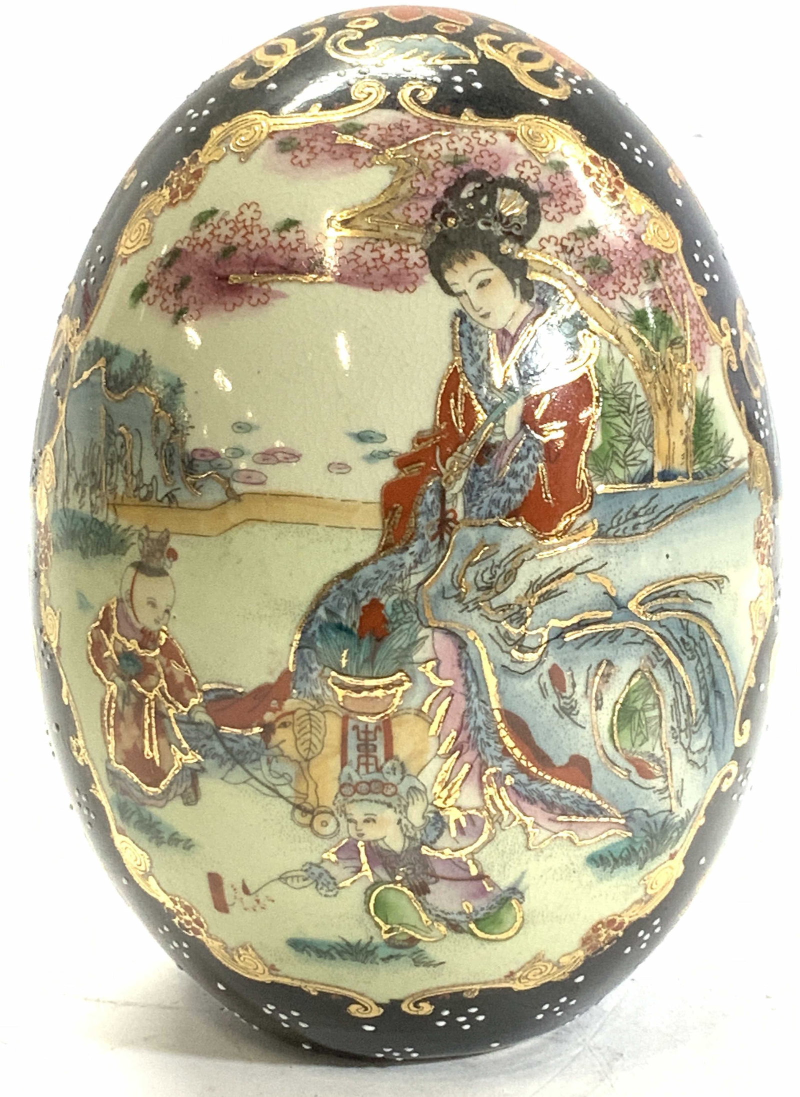 Chinese Hand Painted Porcelain Egg: Porcelain egg hand painted , black and gilt with images of a lady and children in a garden sitting near a cherry blossom tree with mountains in the background. Measures 6.25 x 4.5 in. Asian porcelain