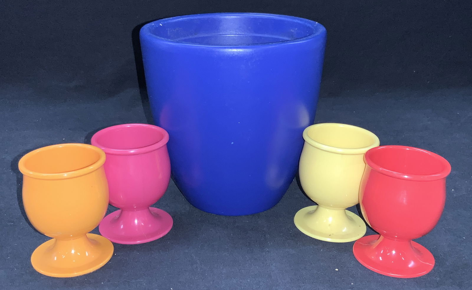 Set 5 Ceramic Vase & Egg Cups: Set of home accessories. 1 Blue toned ceramic vase marked made In Germany on underside and 4 plastic colored egg cups, yellow, red, orange and pink, approximately 2.75 inches tall. Moderate wearing to
