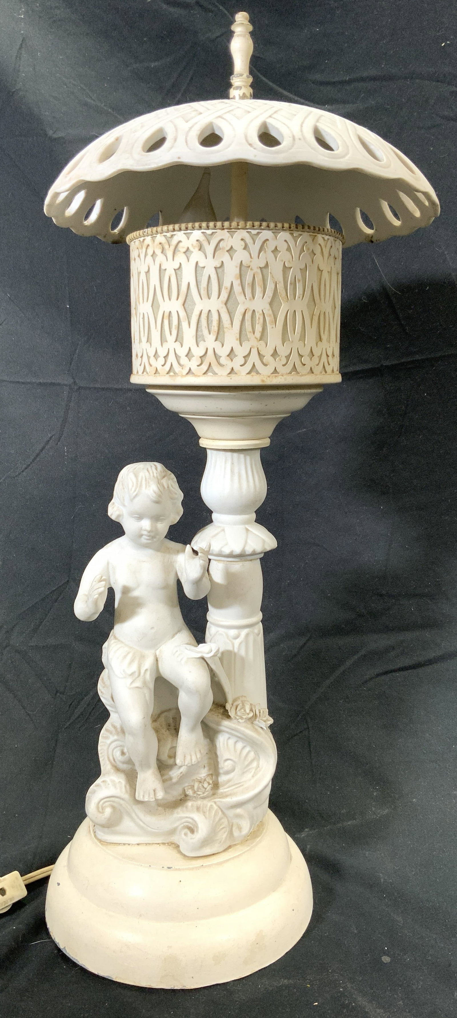 Vintage Porcelain & Metal Cherub Lamp: Lamp is vintage possibly antique. Piece is white toned, porcelain & metal. Piece has umbrella style shade with pierced detail. Base has seated cherub figure. Lamp measures approx 20 inches tall 9 inch