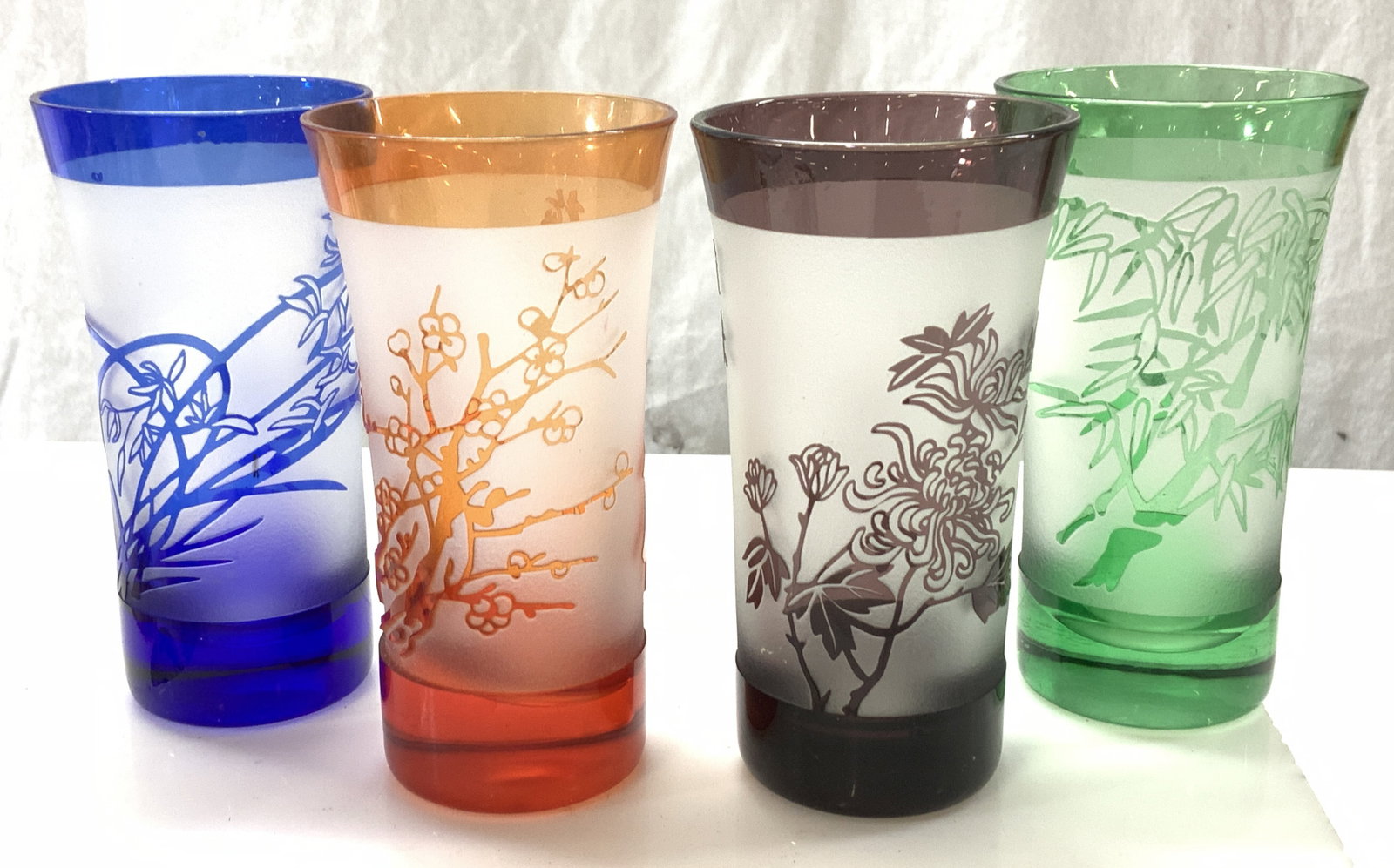 Set 4 Colored Glass Asian High Ball Glasses: 4 signed High ball glasses with Asian florals and Asian characters in colors of amethyst, Orange, Cobalt blue, and emerald , 4.25 x 2 in. Partially with frosted glass . Exterior rim chip on orange. As