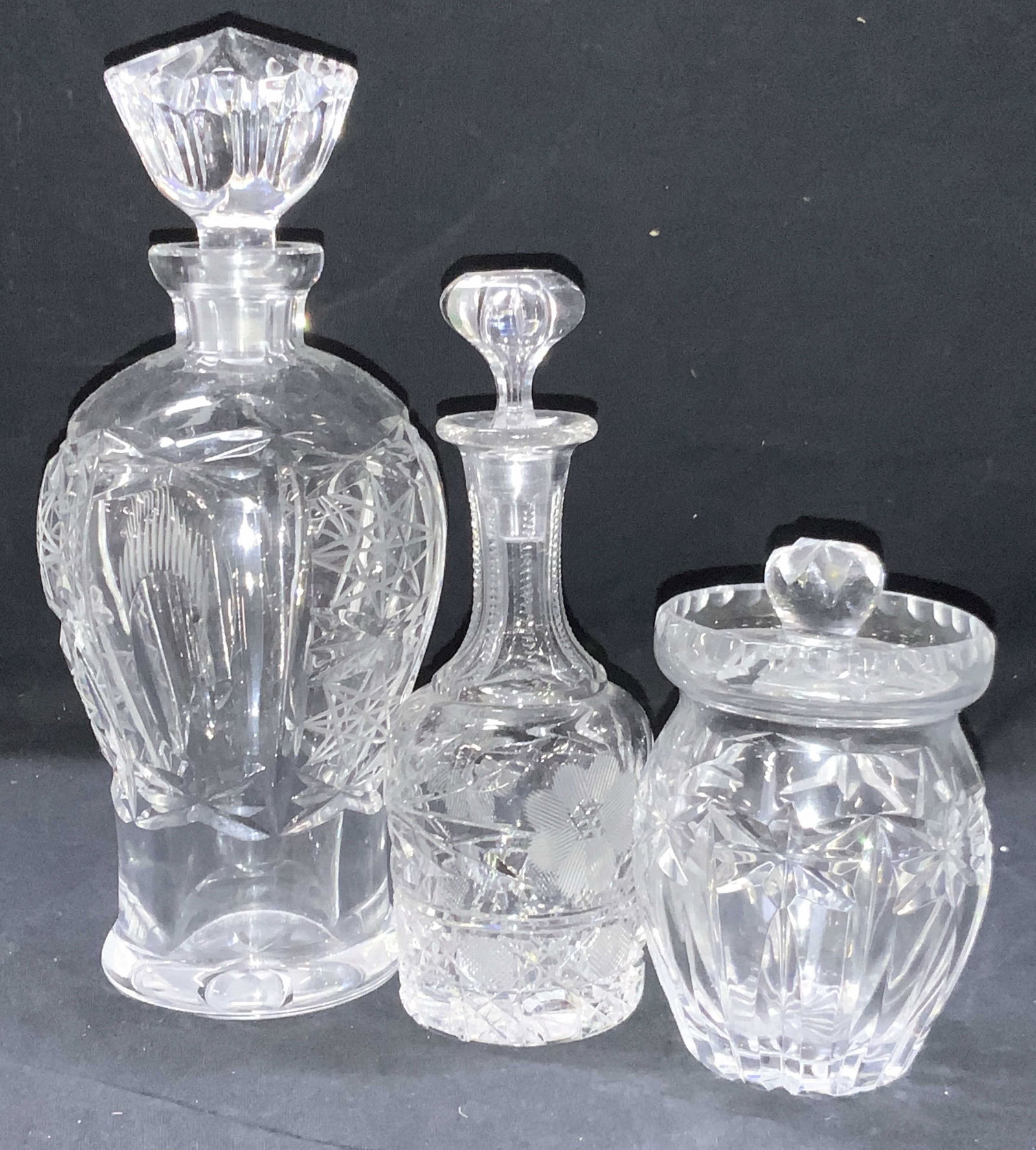 3 Cut Glass Decanters & Jars: 3 cut glass decanter and jars. 1 honey jar with starburst cutouts on exterior. 1 decanter with narrow neck with floral cut motifs and 1 taller decanter with etchings of star patterns. Tallest decanter