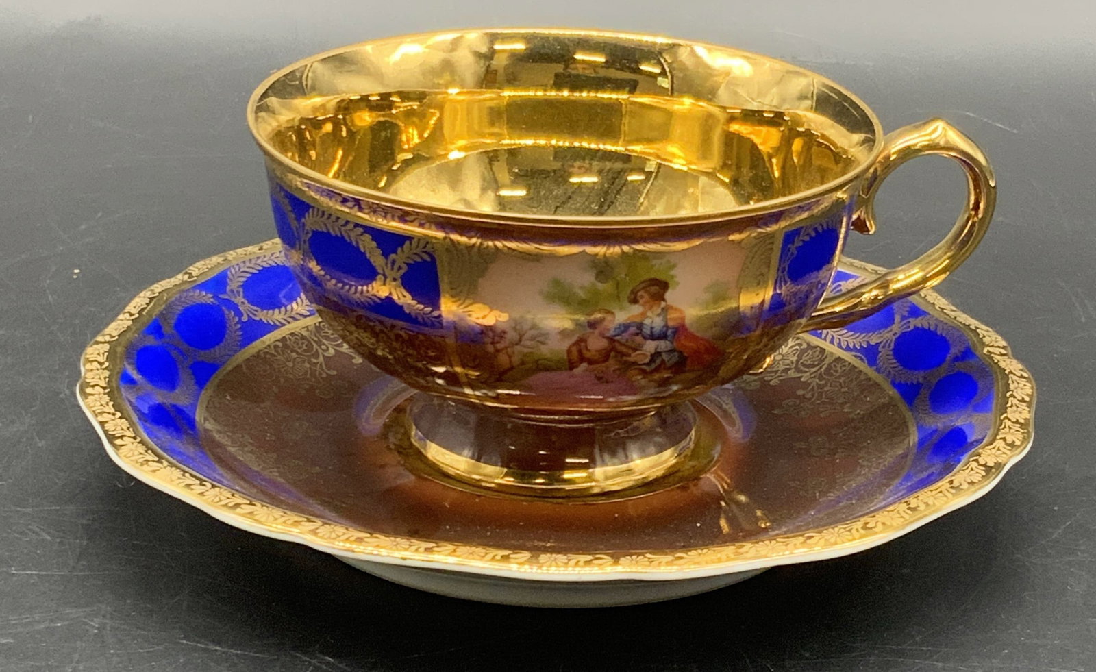 JOHANN HAVILAND BAVARIA Porcelain Teacup & Saucer: A vintage teacup and saucer. Primarily cobalt and brown hues decorated with gilt line work. Interior of the teacup is gilt lined. Vignettes featuring men and women in nature to the obverse of both the