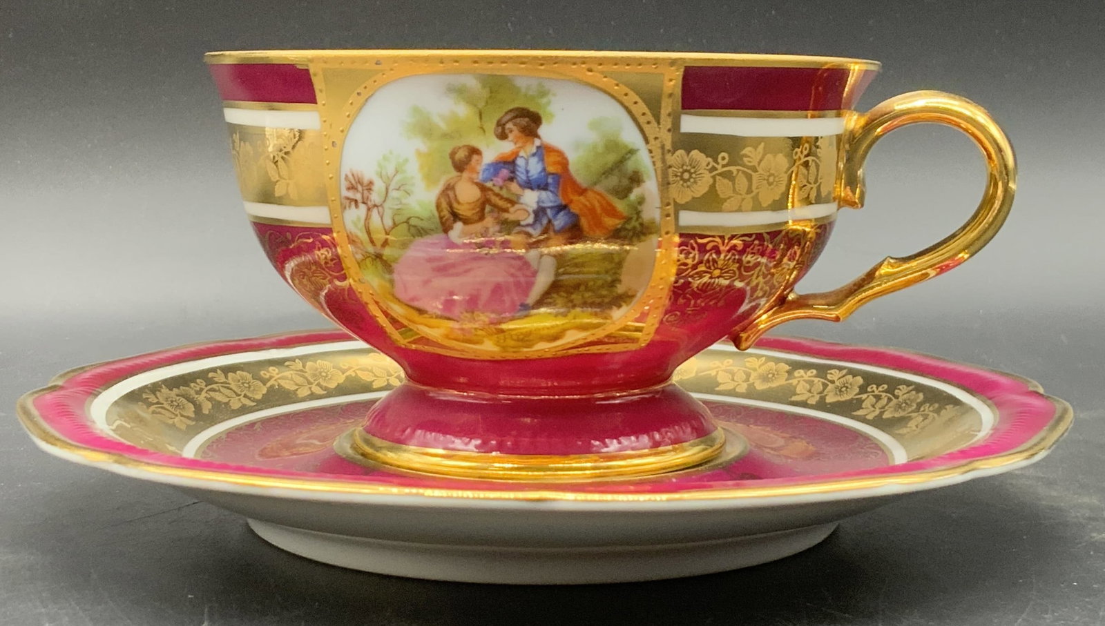 HUTSCHENREUTHER Vintage Porcelain Teacup & Saucer: A vintage teacup and saucer. Primarily raspberry hued decorated with gilt line work. Interior of the teacup is gilt lined. Vignettes featuring men and women in nature to the obverse of both the teacup