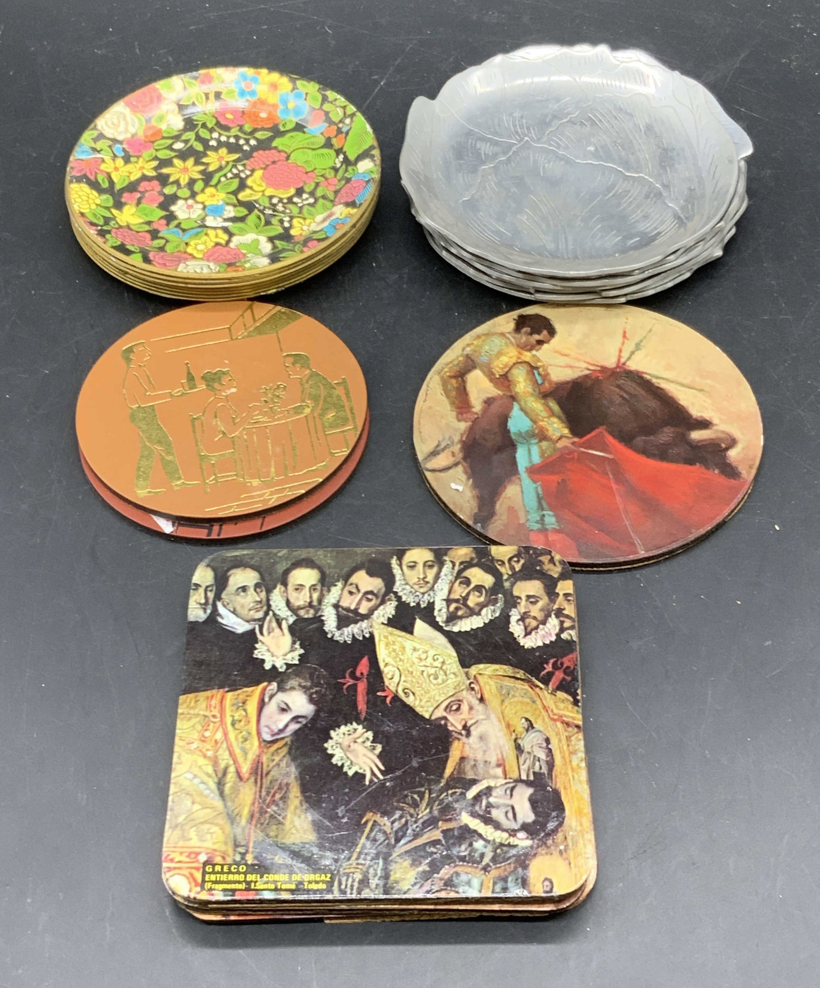 Group Lot 20 Coasters, HOME LINES & More: A group lot of coasters crafted from metal, wood, cork, or composite materials. Coasters are circular, square, or leaf forms. Brands include HOME LINES, GRECO Entierro Del Conde De Orgaz, and more. Pi