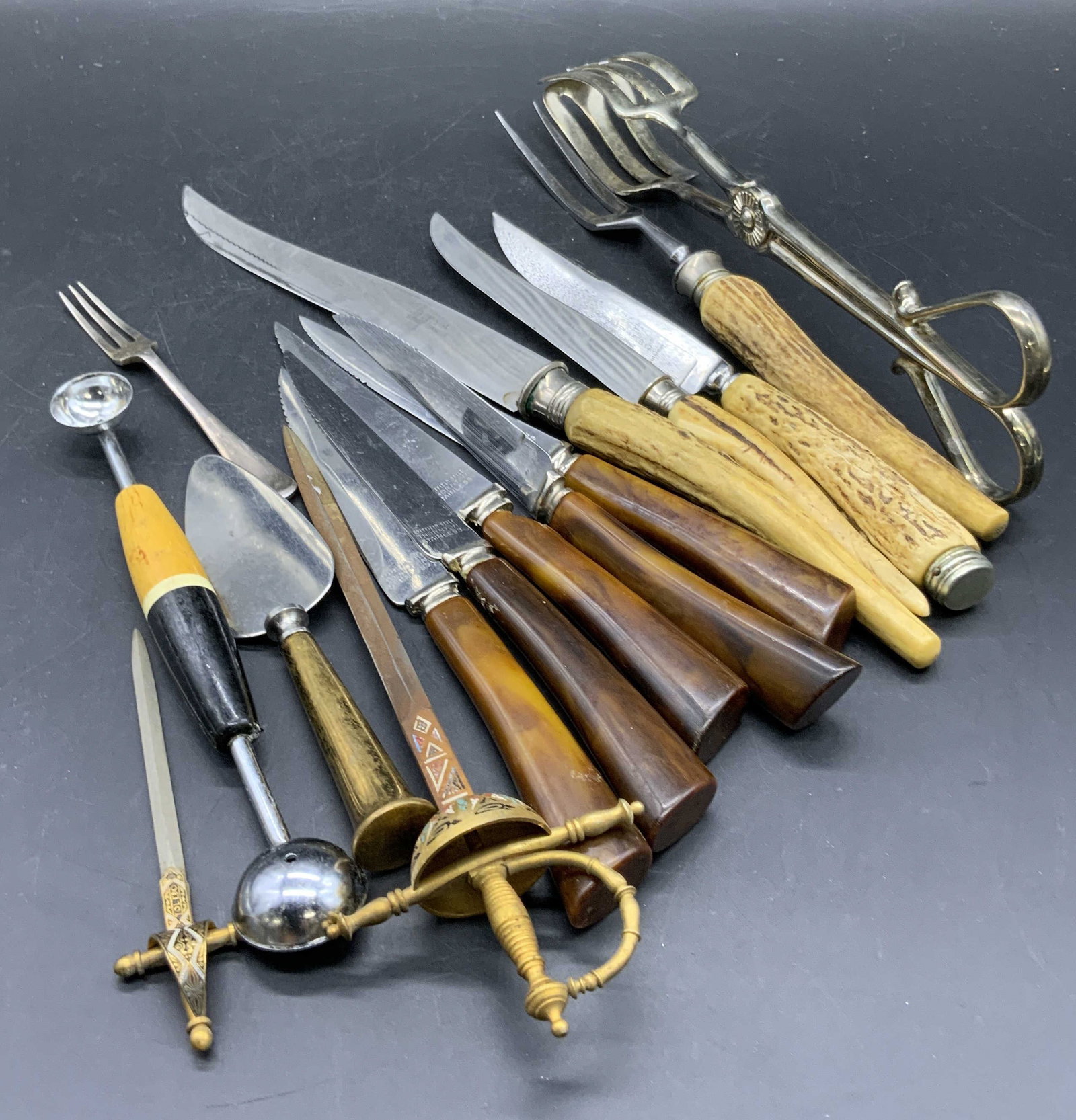 Lot14 Vintage Utensils, Horn Handled Cutlery &more: A group lot of vintage utensils / cutlery. Pieces include knives, melon baller, tongs, serving utensils, letter openers, and more. Tongs measure approx. 10L x 2.5W in. Measured at widest point. Brands