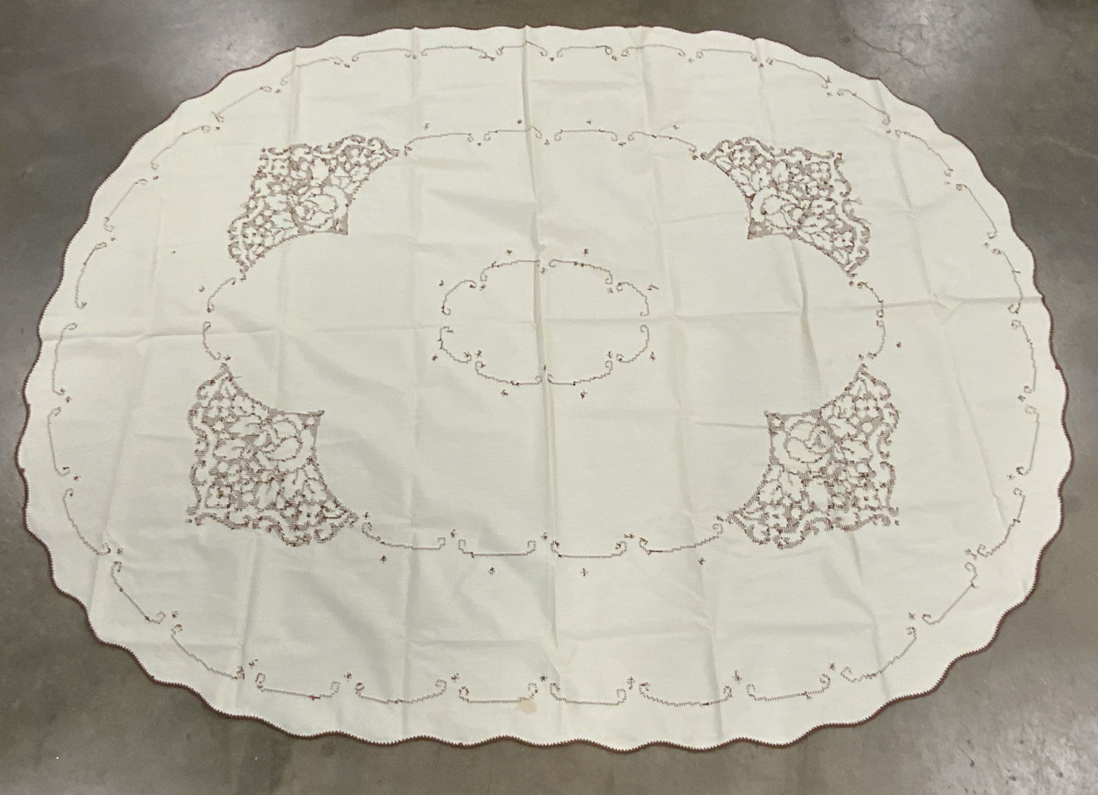 Vintage Scalloped Rim Embroidered Tablecloth: A vintage cream hued oval from tablecloth with a scalloped rim and brown hued embroidered details throughout. Stitched florals towards the center and needlework to the rim. Measures approx 76L x 58W i