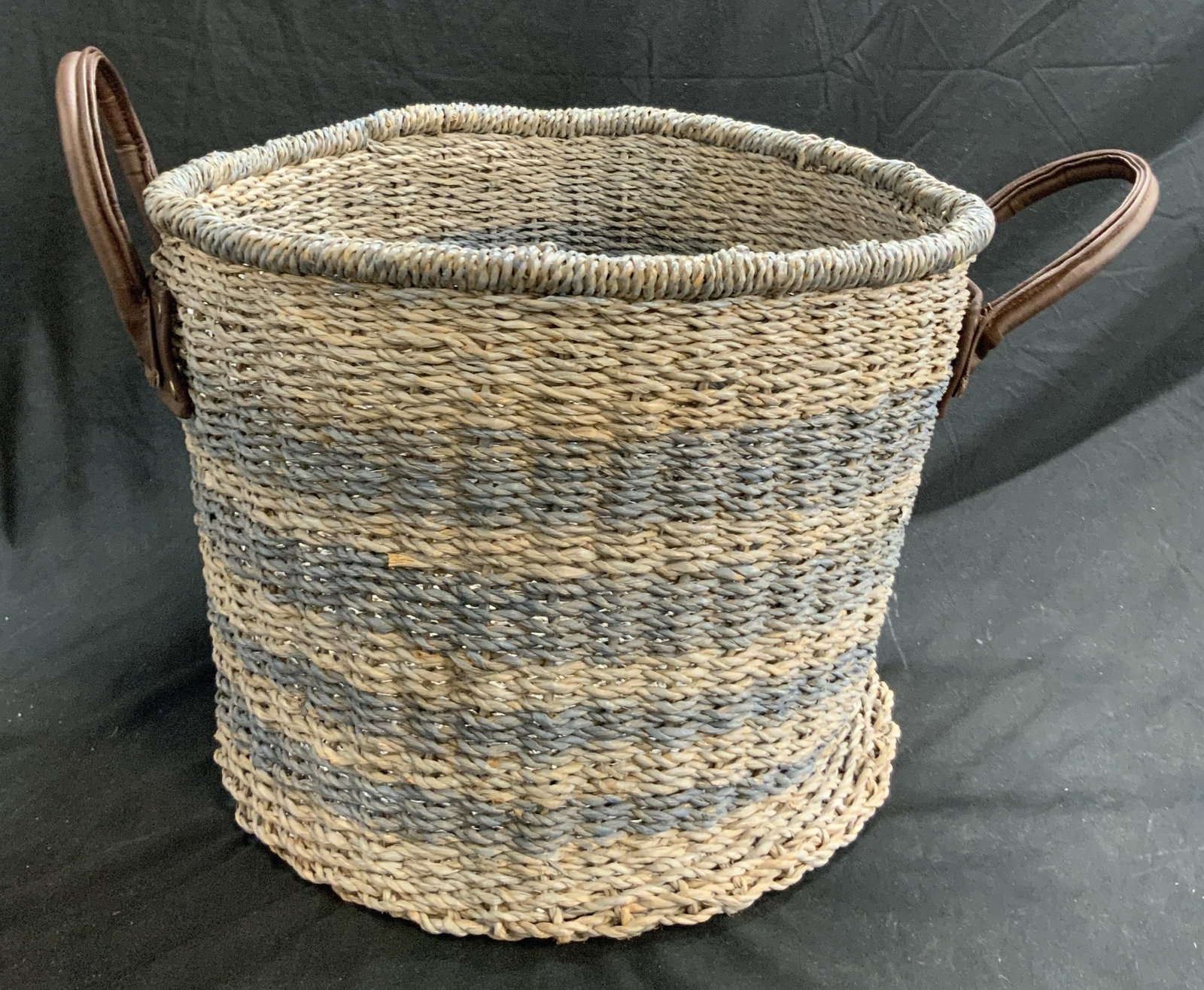 Woven Handled Floor Sized Basket: A basket woven from natural materials with brown hued leather style handles on either side. Basket is striped blue hues. Measures approx. 12H x 17W in. Measurement does not include handles. Some surfa