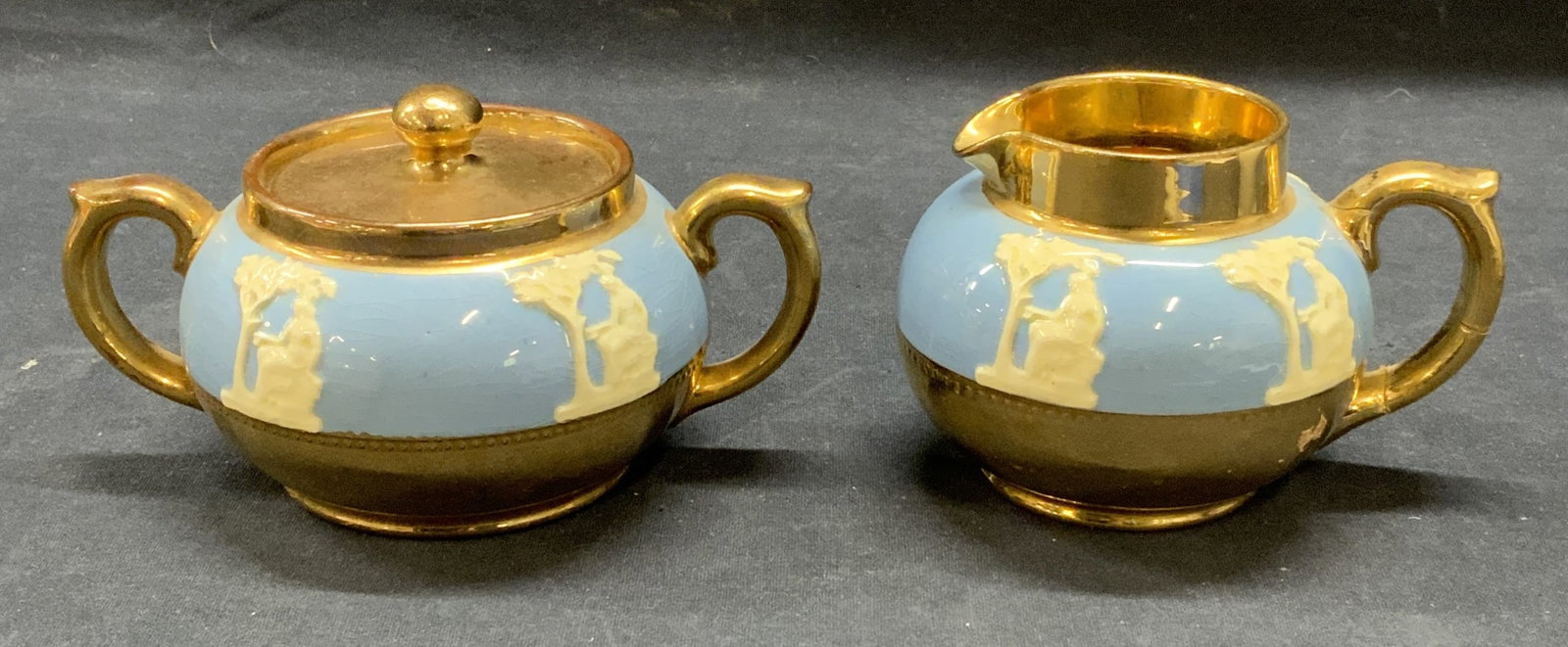 2 Gibsons Gilt Ceramic Creamer & Sugar Bowl: Lot of 2 Gibsons gilt ceramic Wedgwood blue Grecian figural sugar bowl and creamer. Trademarked to undersides. Made in England. Sugar bowl measures approx. 4 x 3 inches. Creamer measures approx. 3.5 x
