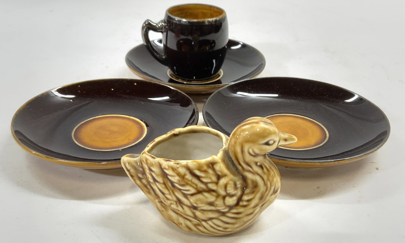 Lot 5 Cer Espresso Cup Saucers & Duck Figure: Lot includes 1 espresso cup, 3 saucers & duck shaped vessel. Each piece appears to be ceramic possibly porcelain. Saucers and cup are brown and goldenrod toned. Saucers measure approximately 4.75 inch