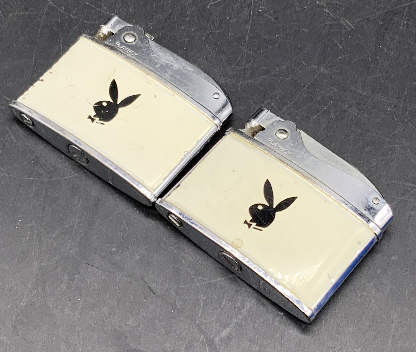 Lot 2 PLAYBOY BUNNY Lighters: Two PLAYBOY BUNNY lighters. Silver, black, and white hues. Iconic bunny motif to the center. Measures approx. 1.75L x 2W in. Some losses present, some surface wear consistent with age and use. Some sc