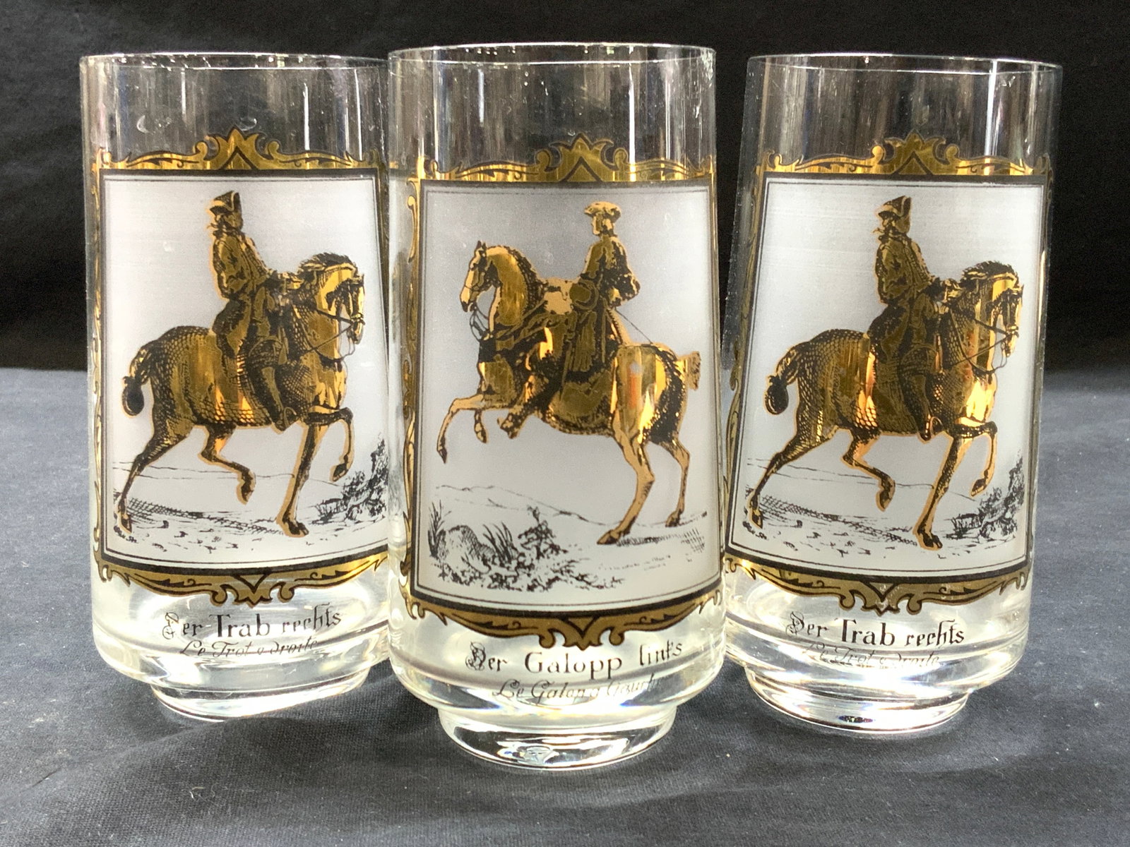 3 Gilt Horse Riding Glassware: Set of 3 glasses with images of men riding horses in white and girl accents. 2 reads in French the trot to the right, and 1 reads the gallop to the left. Approximately 5 inches tall Minor wearing to g
