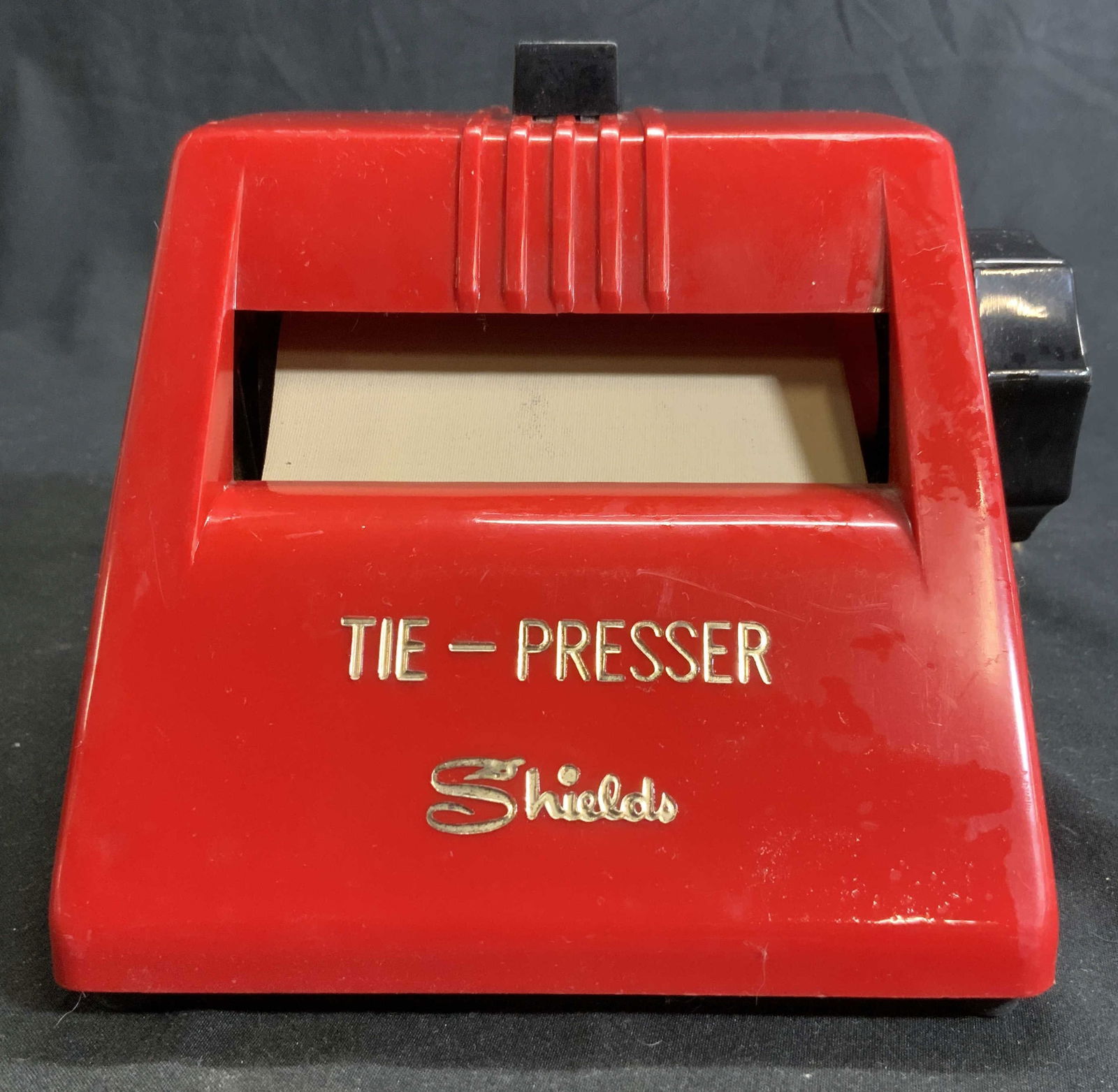 SHIELDS Vintage Tie Presser Machine Red: Front reads SHIELDS TIE PRESSER. Piece is red with gold toned lettering, black toned knob at side & REVERSE switch at top. Piece is plastic possibly Bakelite. Tie presser measures approx 5.25 inches t