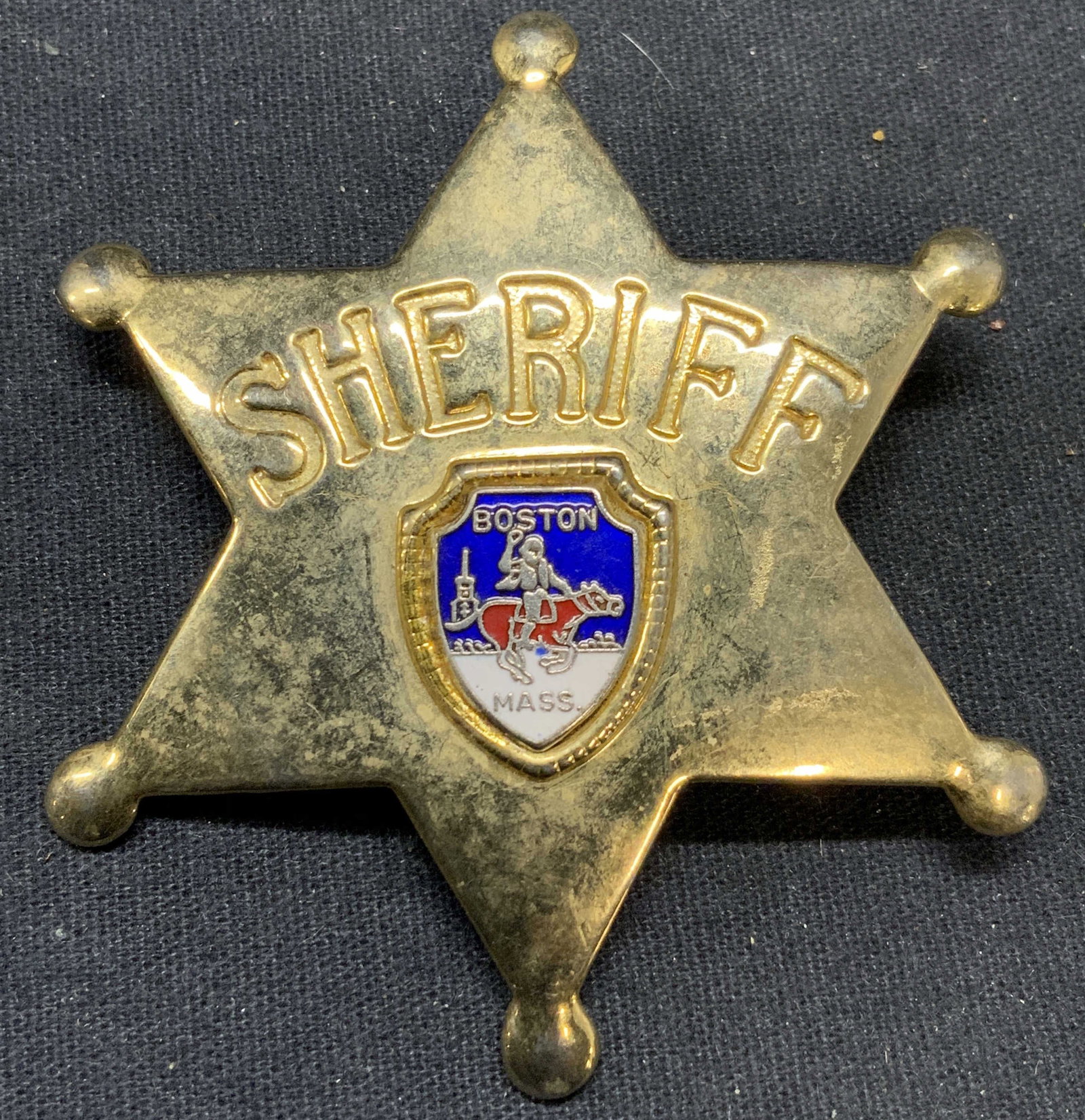 FORT Gold Tn Boston Sheriff Badge: Gold tone and enamel Boston, Massachusetts sheriff badge. Trademarked Fort to underside. Hinge pin to underside. Measures approx. 2.75 inches wide. Some surface wear including scratching observed. Est