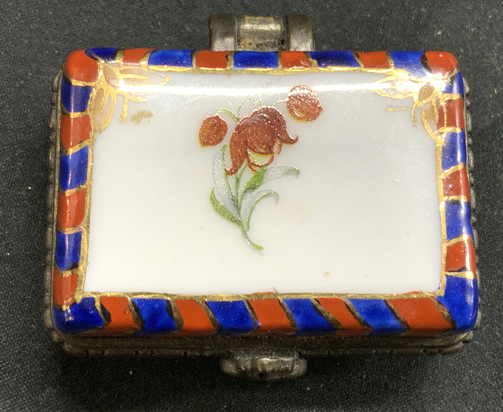 Vintage Hand Painted Porcelain Pill Box: Pill box is vintage. Piece is hand painted and porcelain. Box has red, blue & gilt diagonal stripe detail. Center of lid has floral & leaf detail. Rim & hinge is metal. Measures approx 1.75 inches wid