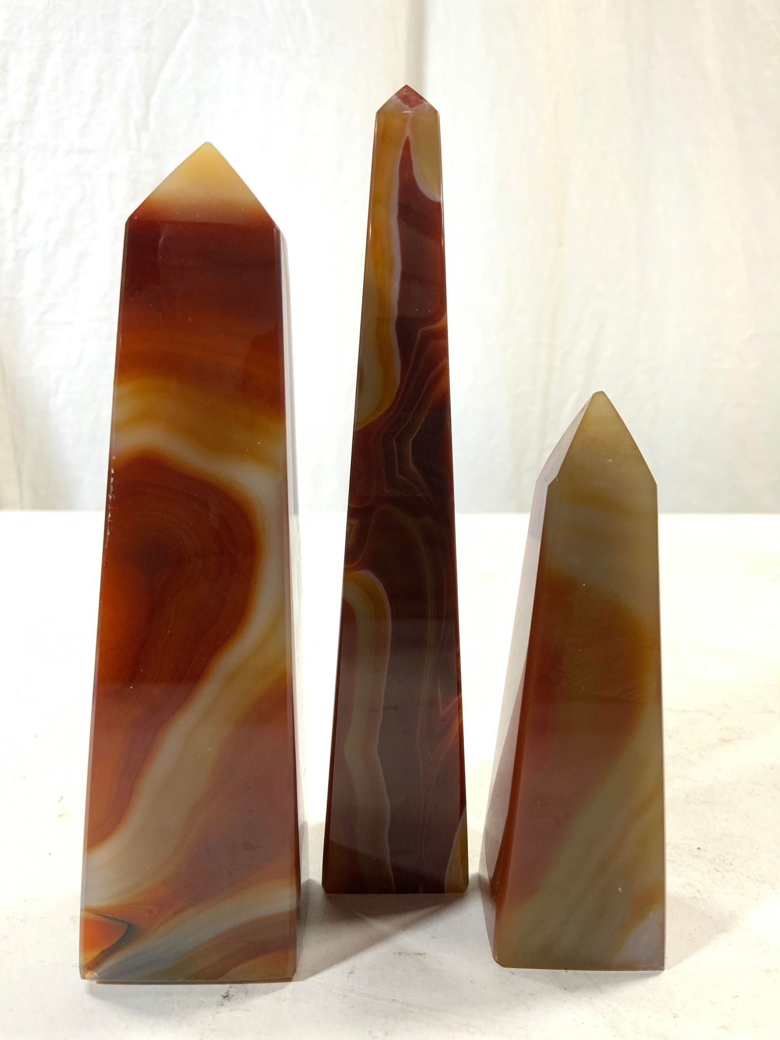 Group Lot 3 Natural Stone Agate Obelisks: Obelisk figures are natural stone appear to be agate. Each have burgundy, maroon & orange toned banding. Largest piece in lot measures approx 6.5 inches tall 1.25 inches square at base. Obelisks, obel