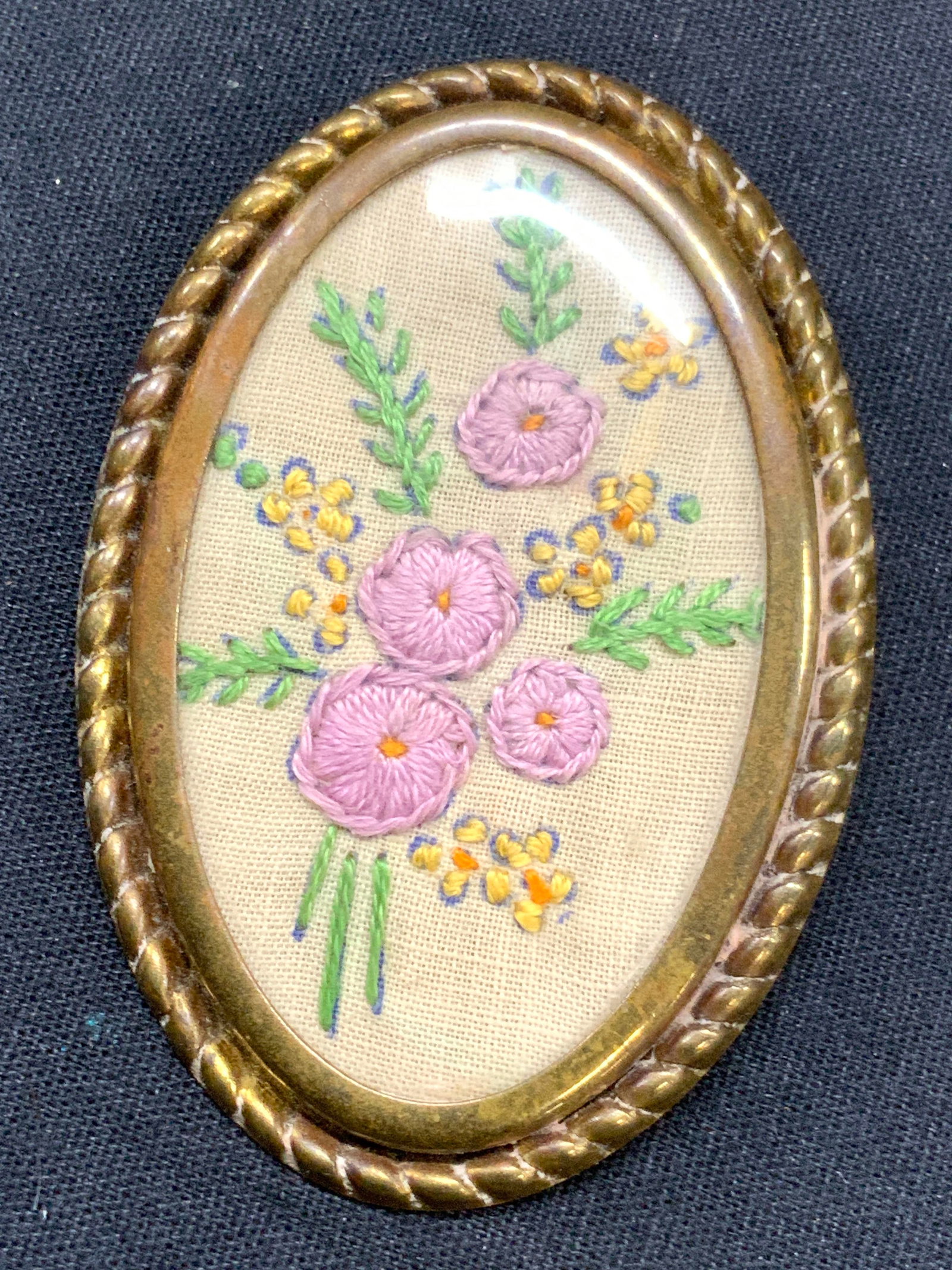 Vintage Brass & Embroidery Brooch: Vintage brooch with brass frame and backing, encasing an embroidered patch with pink flowers and green leaves. Oval shaped with pin backing. Approximately 3 x 2 inches. Evident wearing to backing and 