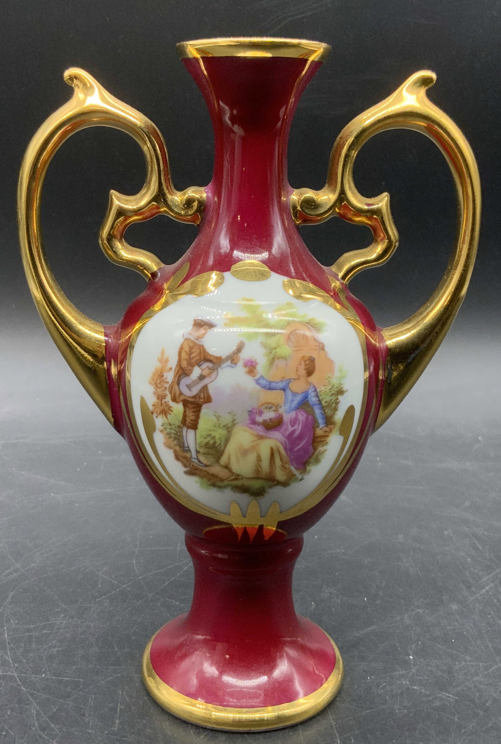 LIMOGES FRANCE Vtg Porcelain Handled Bud Vase: Vintage porcelain bud vase with twin handles on either side. Gilt accents along a red hued background. Center vignette depicts a couple in a nature setting. Underside stamped LIMOGES FRANCE, once in g