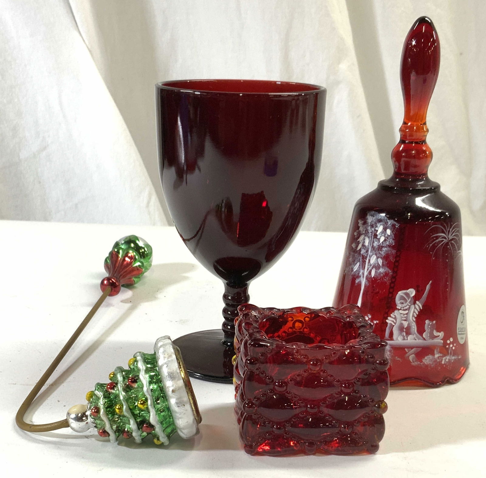 Lot 4 Cranberry Glass Holiday Glassware & Snuffer: Lot includes 1 tagged for FENTON bell, 1 goblet, 1 candle Snuffer & 1 candle holder. Bell has white toned hand painted detail of figure next to tree. Bell measures approximately 6.75 inches tall 3 inc