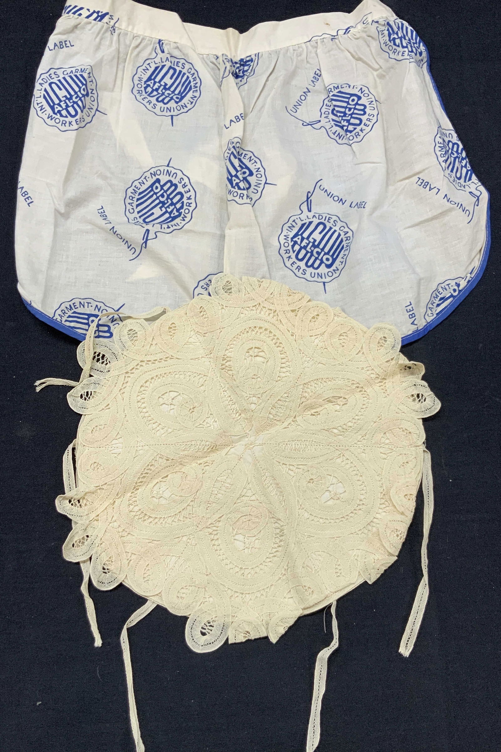 2 Vintage Linen Apron & Lace Roll Keeper: Lot of 2 vintage accessories. 1 of white linen apron with blue throughout reads ladies Garment Union Workers Int. And 1 lace and linen roll keeper. Apron is approx 13 inches wide and 13 inches long wi