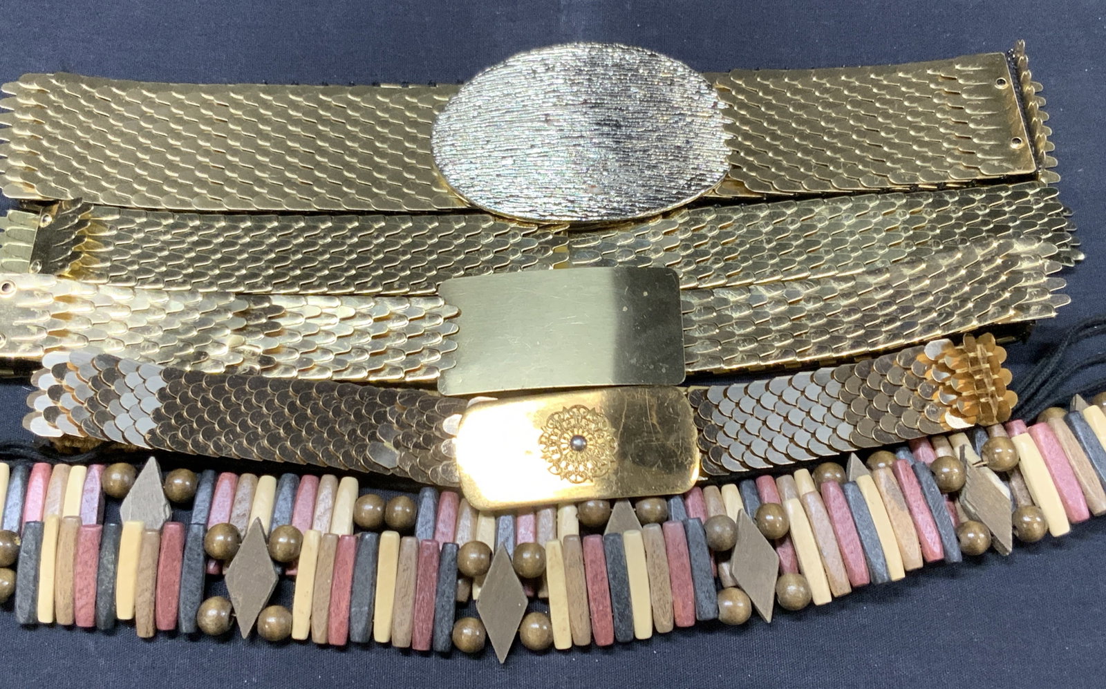 Lot 5 Brass Tn Fish Scale & Wooden Beaded Belts: Lot of 5 belts. 4 made of fish scale style metal with woven fabric undersides. 3 with metal buckles. Lengths range cork 20 inches long to 23 inches long and widest of the 4 belt is 2 inches wide. 1 wo