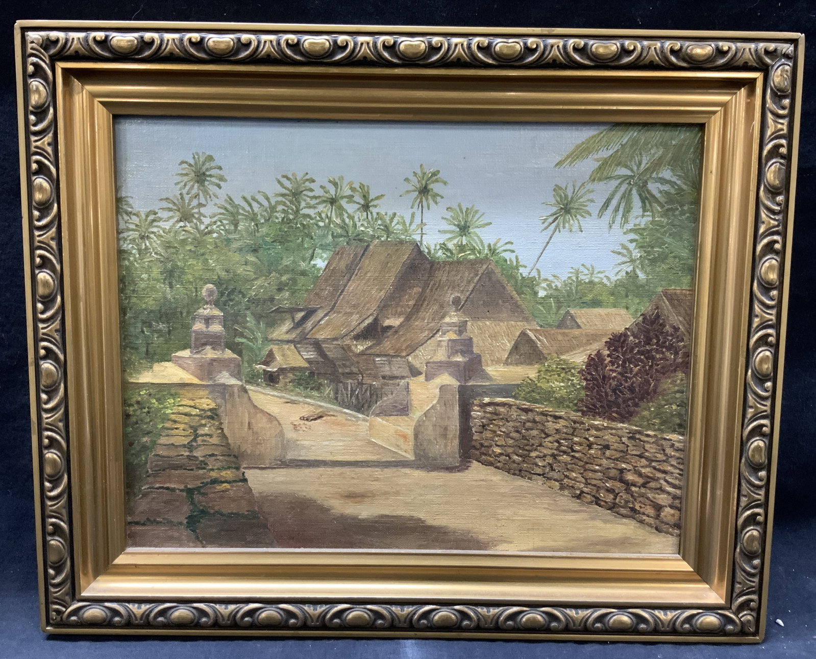 Indonesia Village Entrance Oil Painting: No apparent signature or date Depicts the entrances of an Indonesian village with houses and jungle in the distance. Framed in ornate gold leafed wooden framed with scroll designs. Approx 13 x16 inche