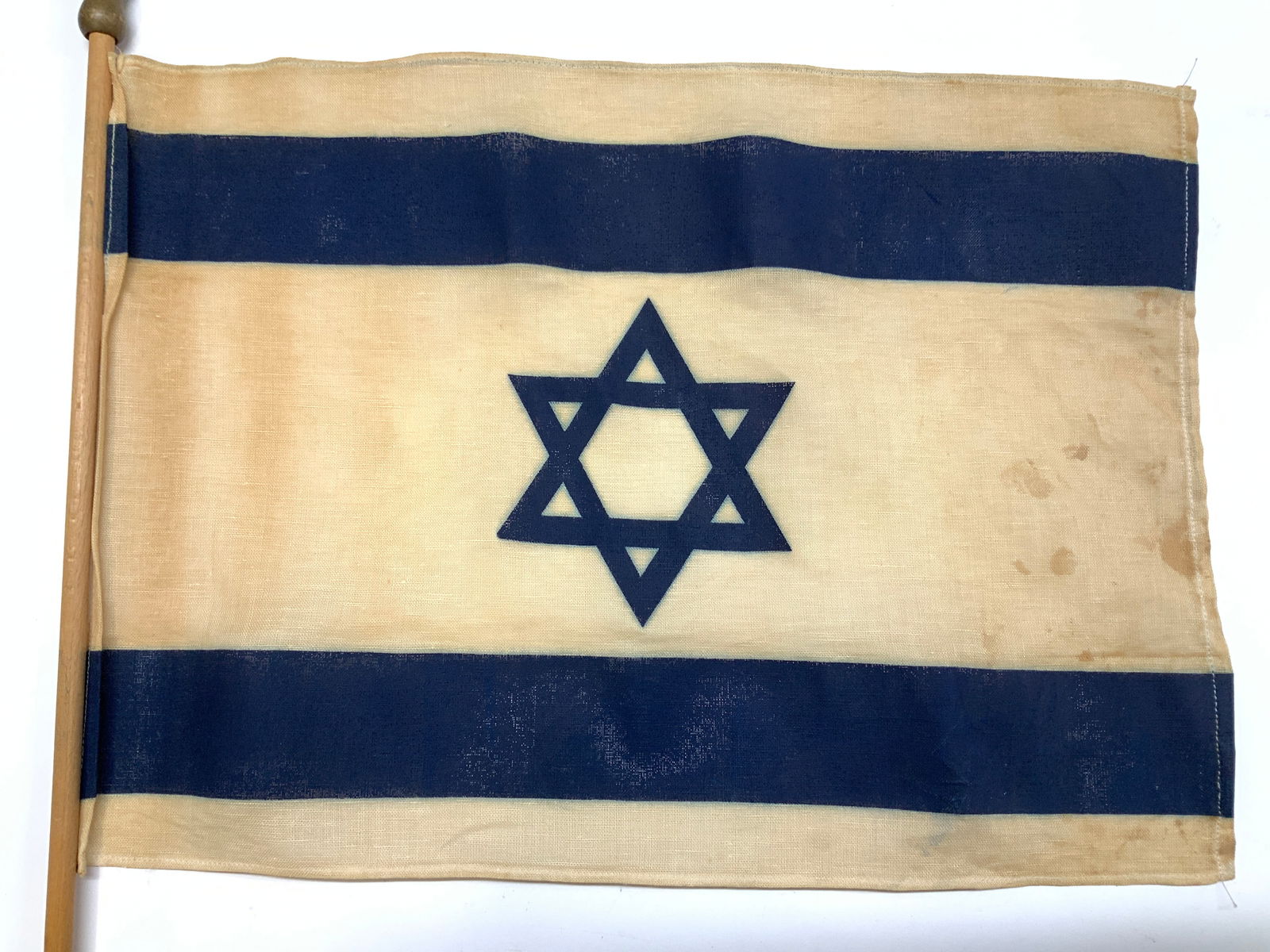 Collectible Israeli Painted Linen Flag: Vintage Hand painted flag of Israel, painted on linen attached to a wooden stick. Height of stick 32 in. Flag measurements 16.25 x 12 in. Yellowing and blemishing to linen, consistent with age, storag