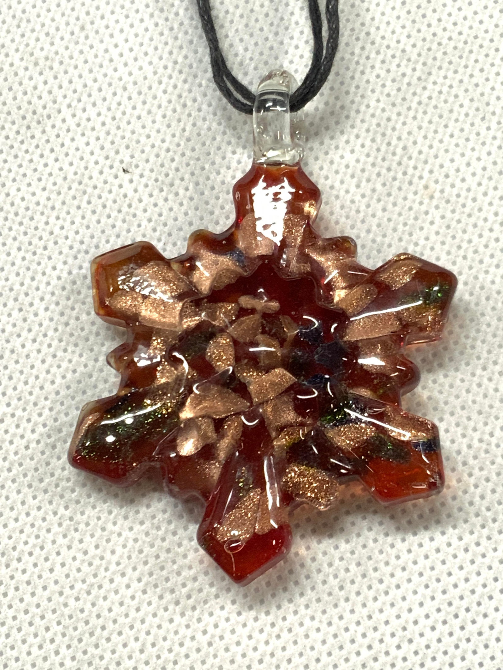 Crafted Glass Snow Flake Necklace: Red toned crafted glass pendant necklace with pendant being in the shape of a snowflake. Affixed to a black cord, approx 8.75 inches long, pendant is approx 2.25 inches long and 1.75 inches wide. Cord