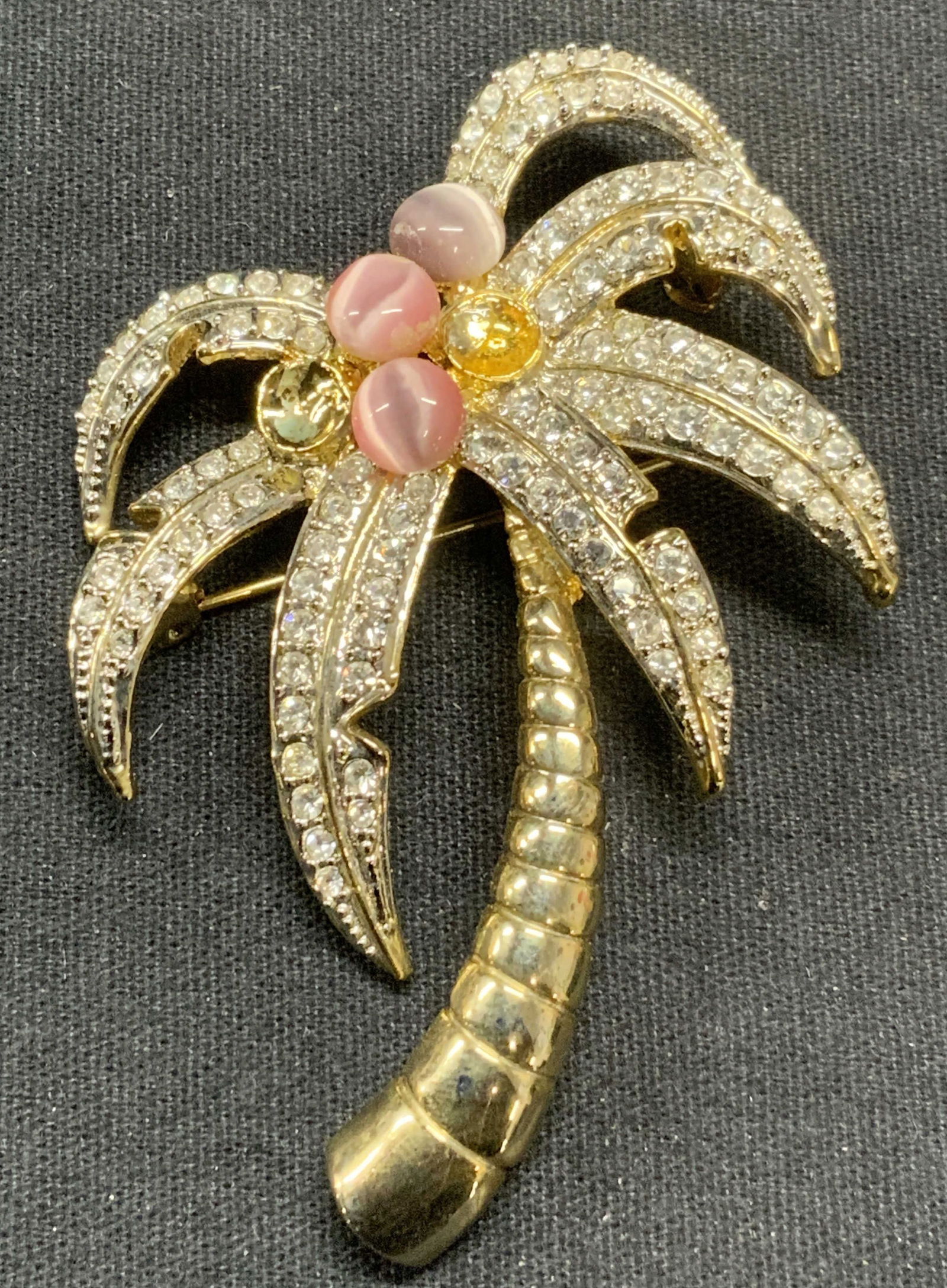 Gold Tone Crystal Coconut Tree Brooch 2.5In: Vintage gold tone coconut tree brooch. Pink toned Lucite Cabochon and colorless crystal decoration. Sculptural three dimensional design. Hinge pin to underside. Measures approx. 2.5 x 2 inches. Surfac