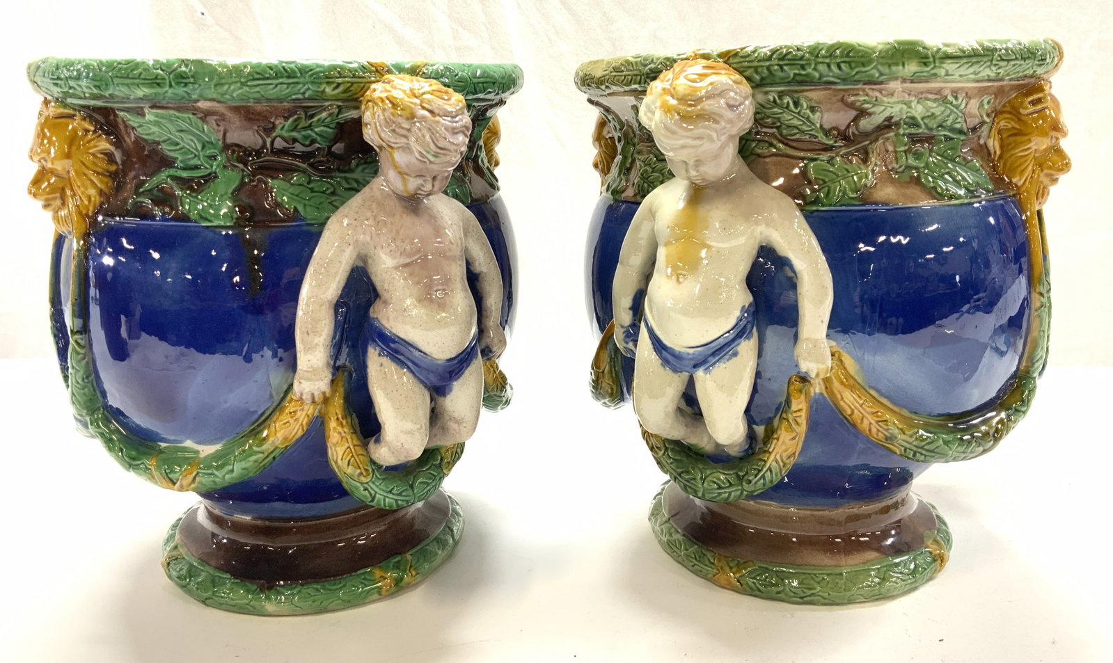 Pr Majolica Jardiniere Cachepot w Putti Handles: These porcelain footed Jardiniere Cachepots are replicas of the antique Mintons Majolica urn Cachepot. Hallmarked with an English Registry Mark ( Diamond with a circle on top), undated. Oversized luxu
