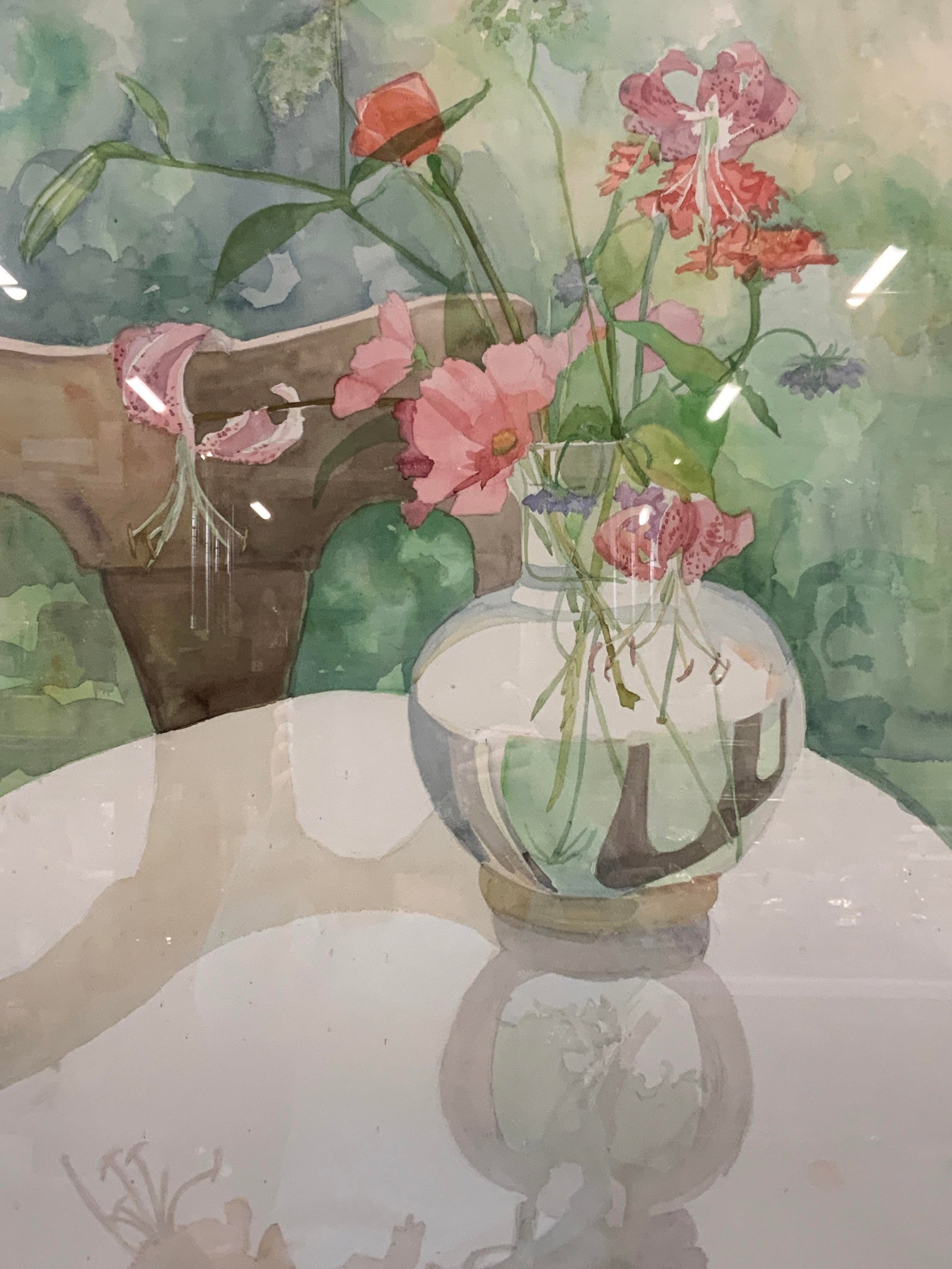 Wendy Shalen Signed Watercolor Still Life 1985 43i: Signed on lower right corner in graphite, 1985 Depicts a vase with pink flowers in water on a table with a wooden chair and shadows on the white table. Framed in a tan toned wooden frame with a green 