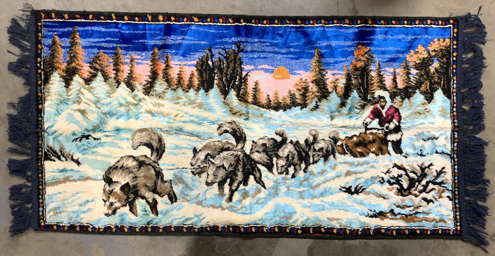 Dog Sled Scene Silk Rug W Fringed Edges: Rug is silk. Piece has landscape oriented dog sled scene. Edges of rug have navy toned fringes. Rug measures approximately 43 inches long with fringes 20 inches wide. Rug, fringed rug, silk rug, dogsl