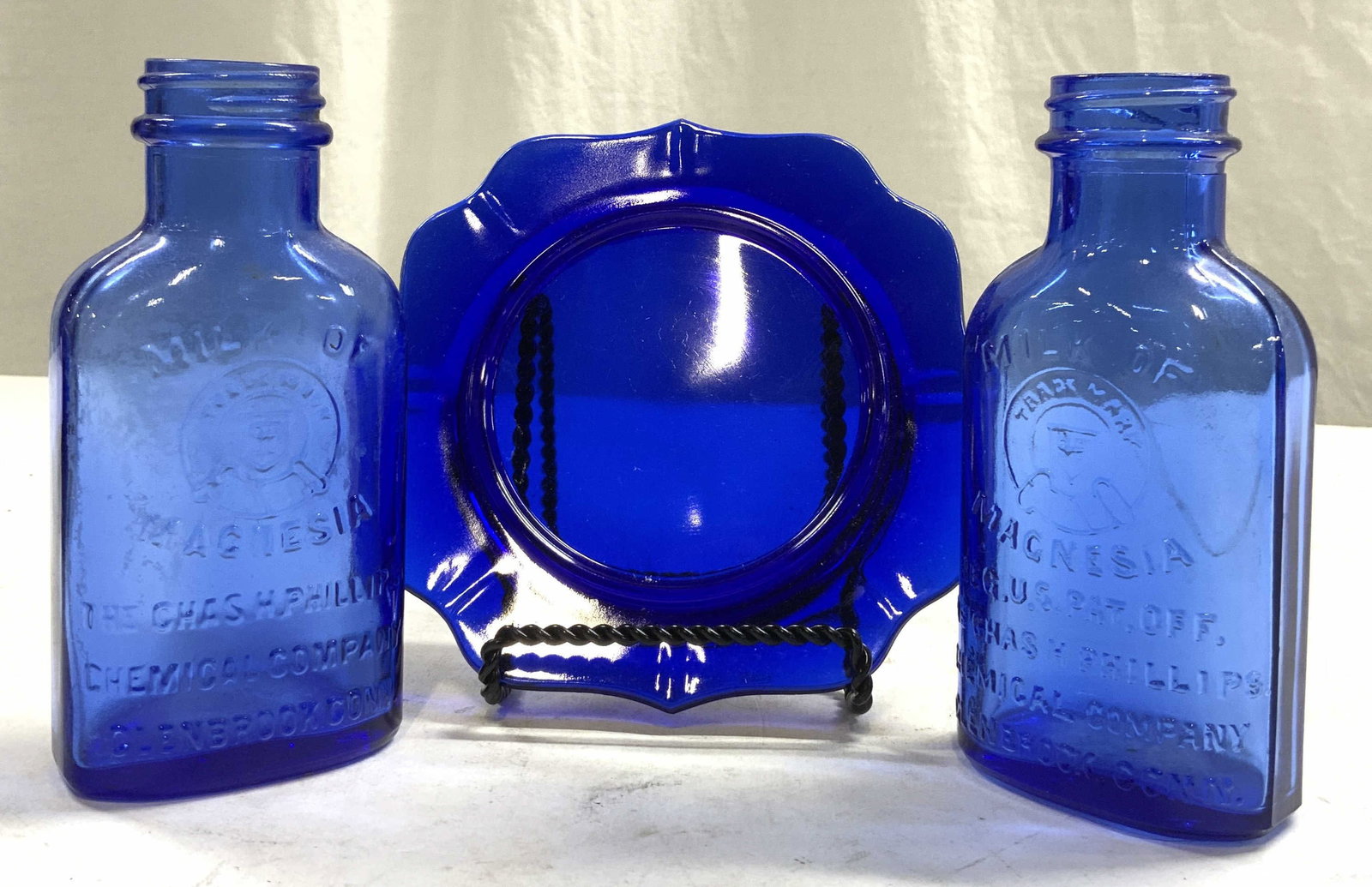 Lot 3 Milk Of Magnesia Glass Bottles & Plate Blue: Lot includes 2 bottles, each read MILK OF MAGNESIA THE CHAS H PHILLIPS CHEMICAL COMPANY, & 1 Art glass plate. Each piece in lot is blue toned and glass. Plate measures approximately 5 inches wide & de