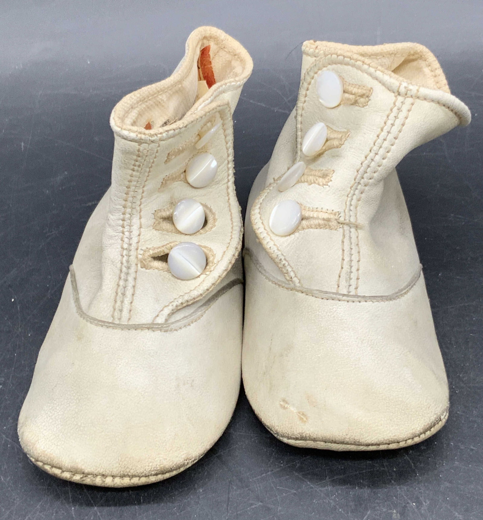 Antique First Shoe Leather Baby Shoes: Antique, possibly vintage, first shoe baby shoes crafted from white hued leather. Pearlescent style buttons to the shoes. Shoes measure approx. 2.5H x 4.25L x 1.75W in. Some paper to interior of shoes