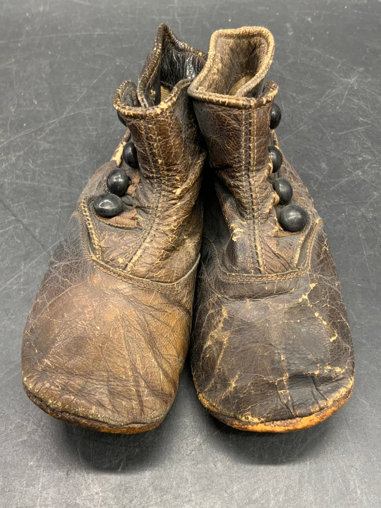 Antique Brown Leather First Shoe Baby Shoes: Antique, possibly vintage, first shoe baby shoes crafted from brown hued leather. Black buttons to the shoes. Shoes measure approx. 2.5H x 4.25L x 1.75W in. Some paper to interior of shoes. Discolorat