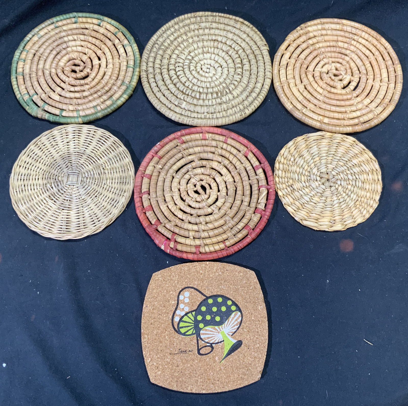 Lot 7 Wicker Rush & Cork Trivets: Lot includes 7 trivets, 1 cork, 2 wicker & 4 rush. Most trivets in lot have circular shape. Cork trivet has signed multi toned painted mushroom detail signed JANE R. Largest trivet in lot measures app