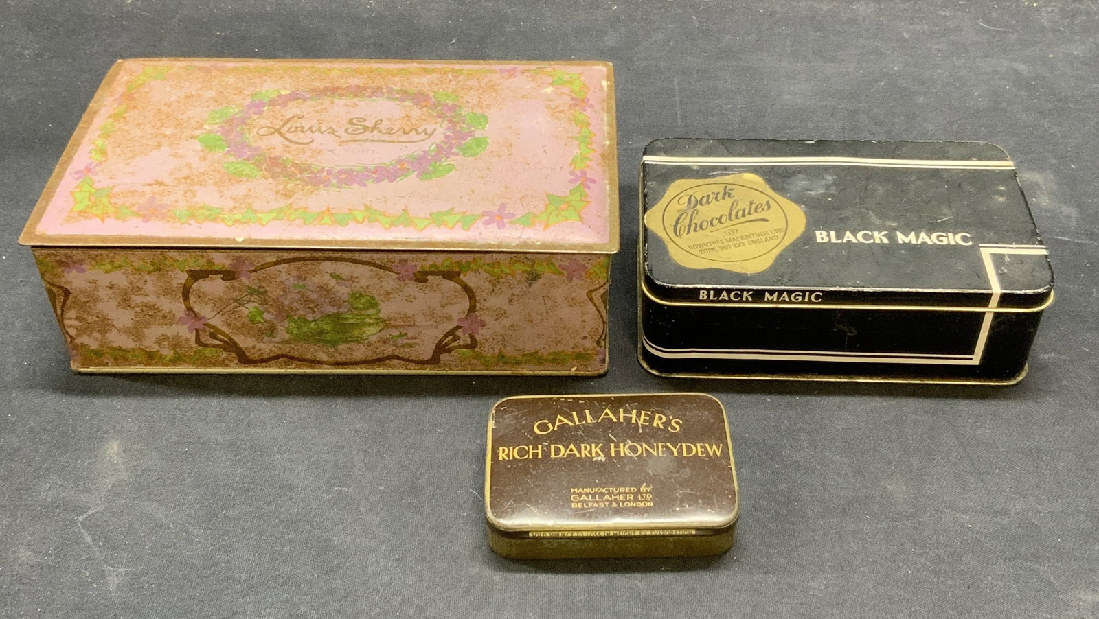 Lot 3 Vintage Tins, Chocolate, Louis Sherry, More: Lot of 3 vintage tins. Gallahers Rich Dark Honeydew, Louis Sherry, and Black Magic Dark Chocolate. Largest tin approx. 8 x 4.75 x 2.5 inches. All pieces have surface wear including scraping and loss t