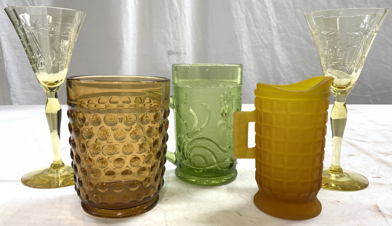 Lot 5 Art Glass Vessels: Lot includes 1 amber toned frosted pitcher, 1 green mug, 2 cordial glasses, & 1 hobnail glass vessel. Cordial glasses each have floral motif relief detail. Cordial glasses each measure approx 6 inches