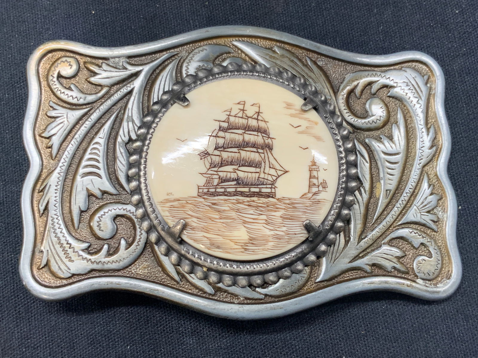Vtg Silver Plate Scrimshaw Maritime Belt Buckle: Vintage silver plate over metal belt buckle with center ornament of carved scrimshaw with etching of a ship on the ocean. Approx 3.5 x 2.5 inches. Scrimshaw measures 1.5 x 1.5 inches. Moderate wearing