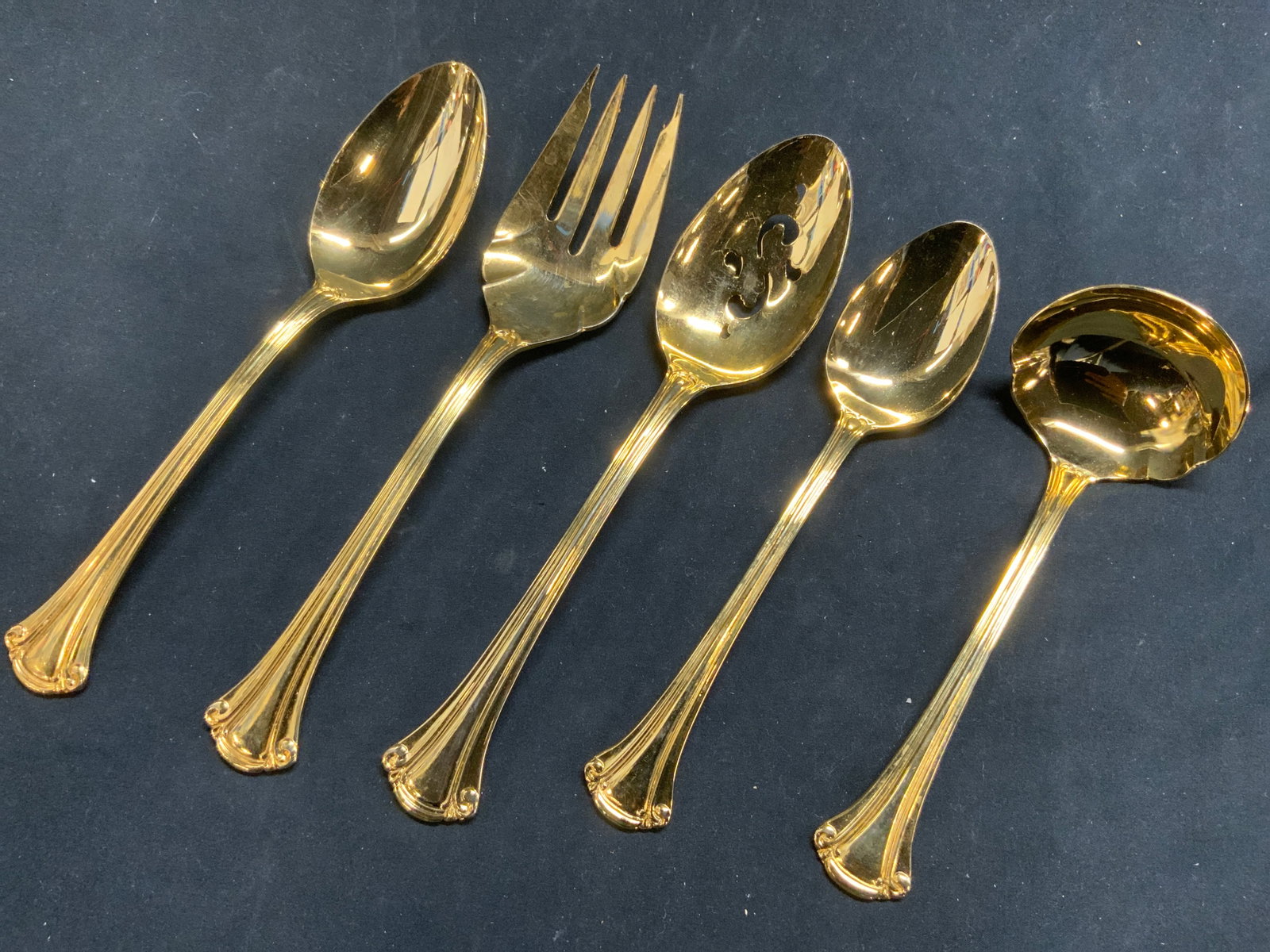 Lot 5 Gold Plated Flatware: Lot includes 2 spoon, 1 serving spoon with pierced detail, 1 serving fork, & 1 ladle. Each piece is gold toned, possibly gold plated. Largest piece in lot (serving fork) measures approximately 8.75 in