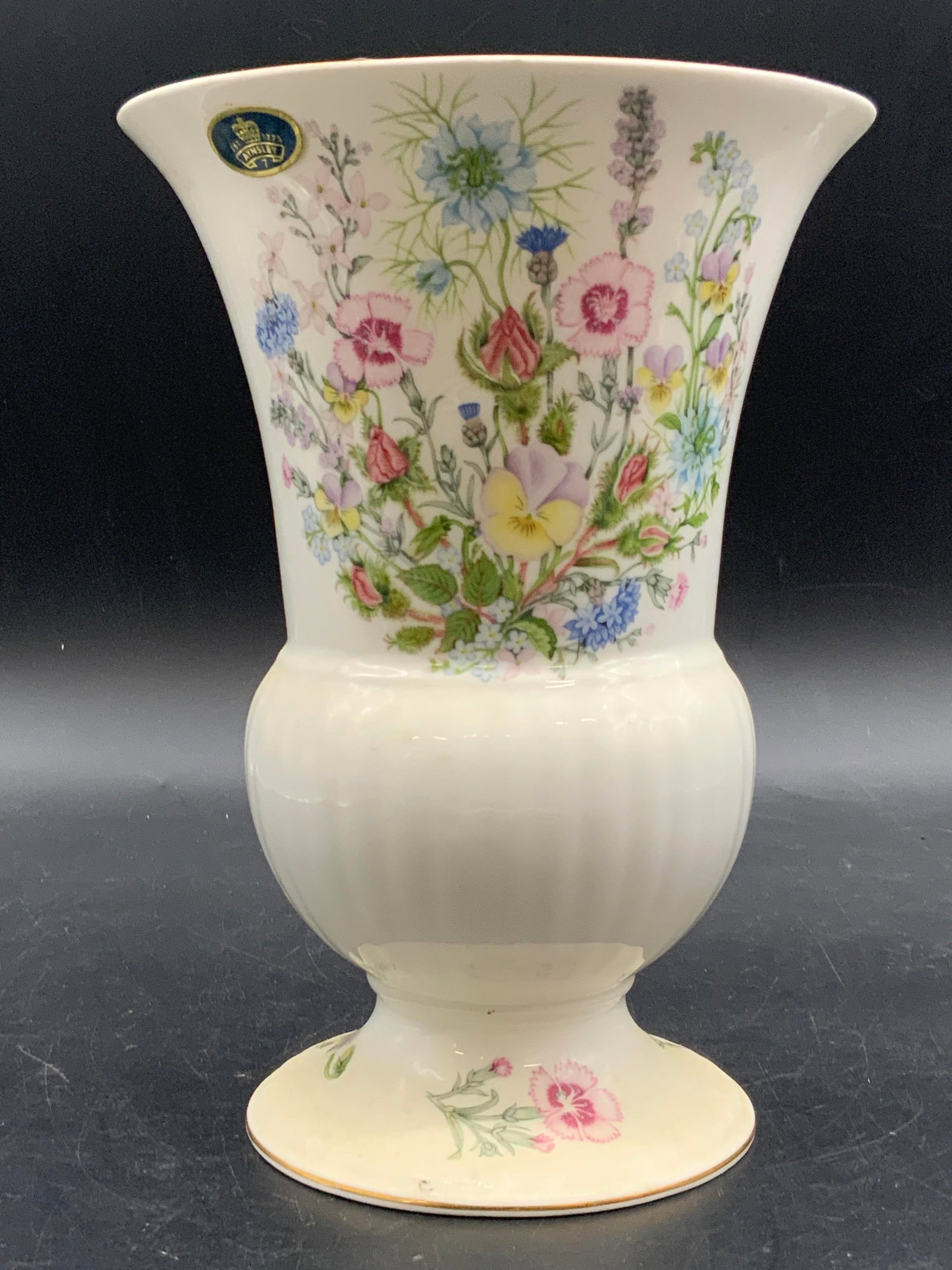 AYNSLEY Wild Tudor English Bone China Vase: A bone china vase featuring a multicolored floral motif. Flared rim with ribbed design towards the base. Measures approx. 7.75H x 5.5W in. Width measured at rim. Sticker towards rim reads AYNSLEY. Und