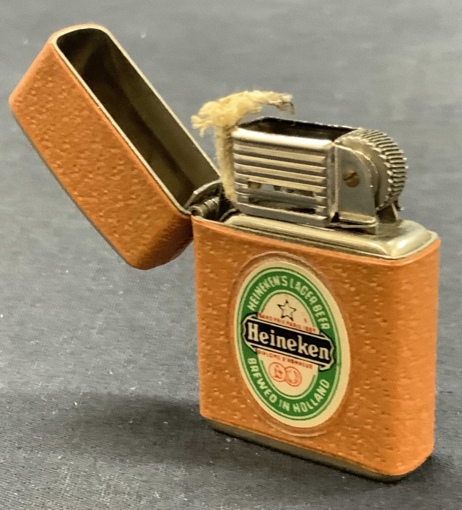 Vintage Heineken Leather Wrapped Metal Lighter: Vintage metal lighter with wick, lined with leather, possibly faux leather and with Heineken logo on both sides Approx 2.25 x 1.5 inches Moderate wearing to metal and leather to lining. Not tested for