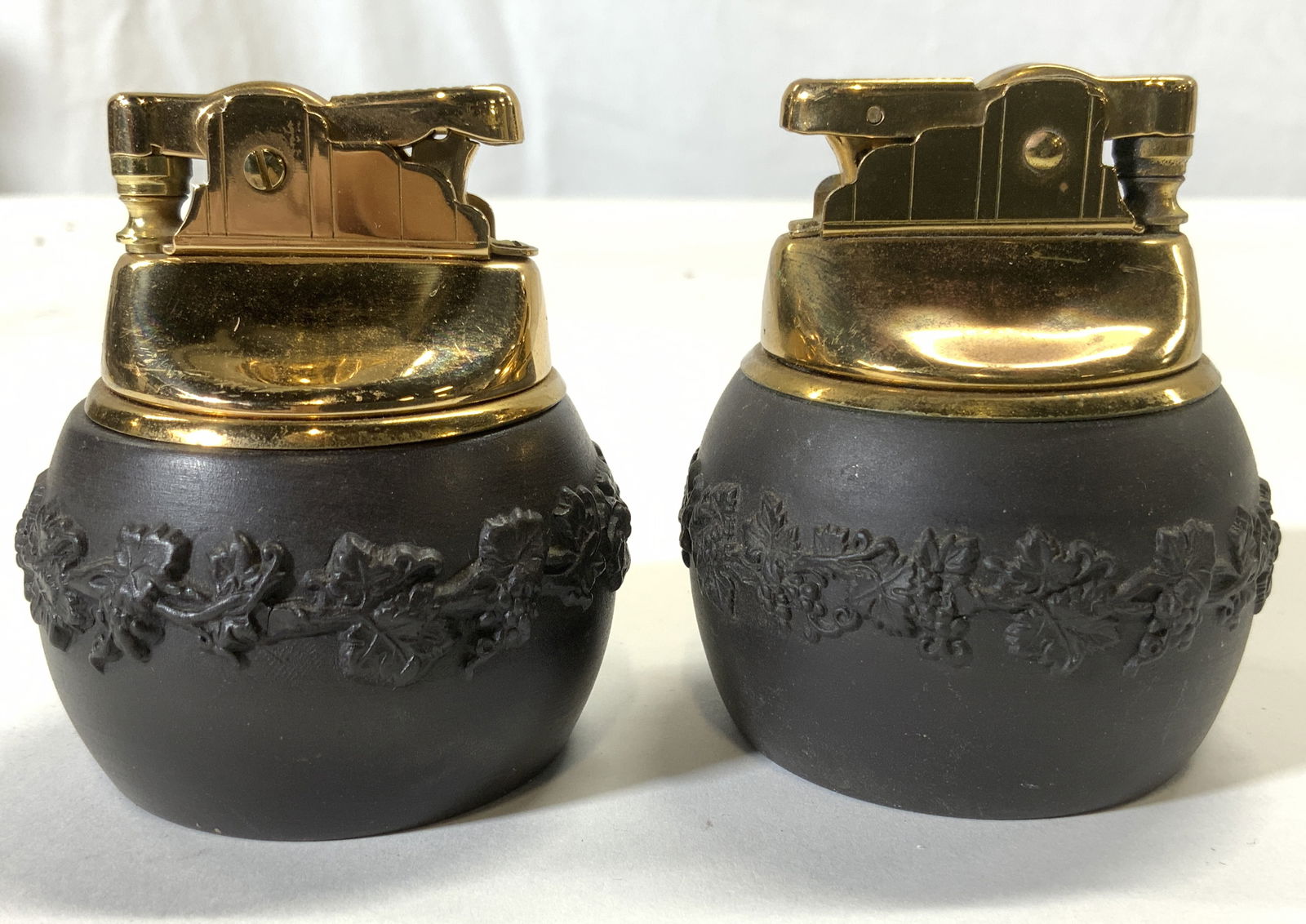 WEDGWOOD Pair Vintage Lighters Black & Gold: Undersides are inscribed WEDGWOOD ENGLAND. Each are black and gold toned. Each have beveled grape leaf & vine detail. Gold toned lighters sit inside black toned Jasperware vessels. Lighters each measu