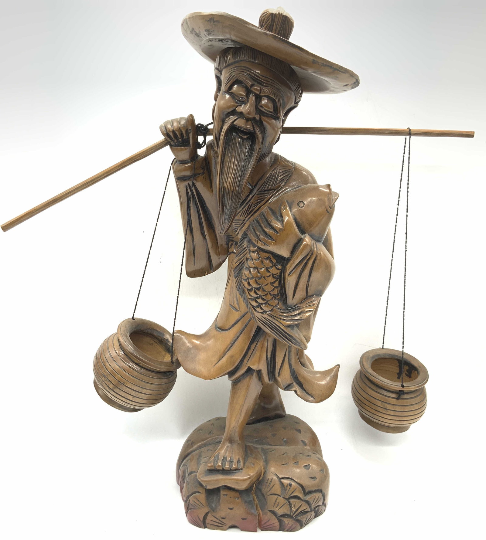 Carved Wooden Asian Fisherman Figural: Carved wooden figure of an Asian fisherman in robes, holding a pole with buckets in his shoulder and a fish in his arms. Approx 18 inches tall and 18 inches wide. Moderate surface wearing to wood thro