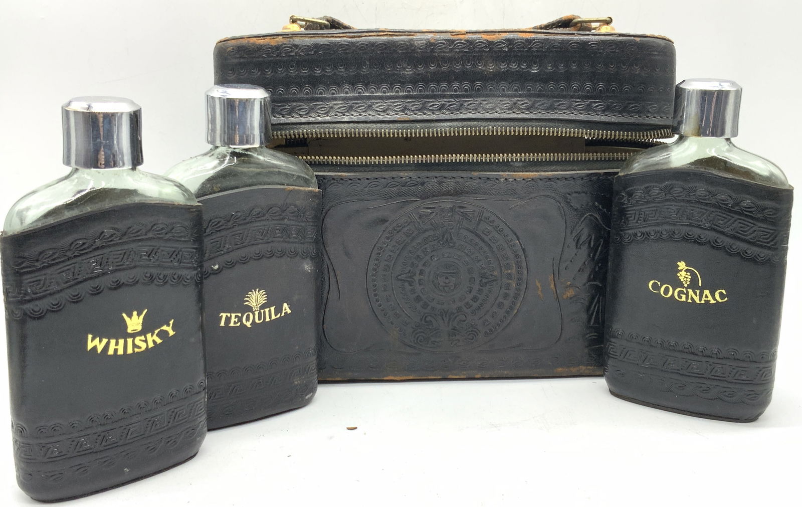 Mexican Leather Flask Travel Carrying Case: Mexican leather flask carrying case with front design resembling Mayan decoration and ornate carvings in animal and geometric patterns. Contains 3 flasks, marked, whiskey, cognac, and Tequila and 2 st