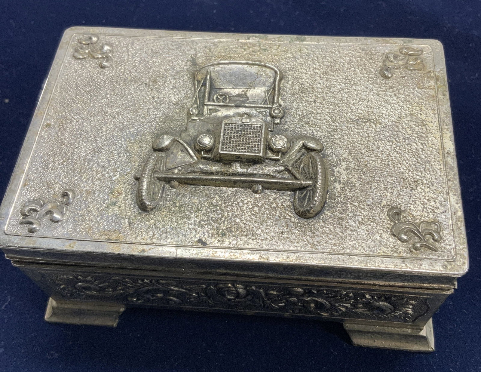 Antique Car Metal Tobacco Box/Trinket Box: Embossed silver tone hinged box. Lid has image of an antique car, side are designed with embossed roses and scrolling. Interior under lid is lined in red velvet. Painted black wood lines the box. Exte