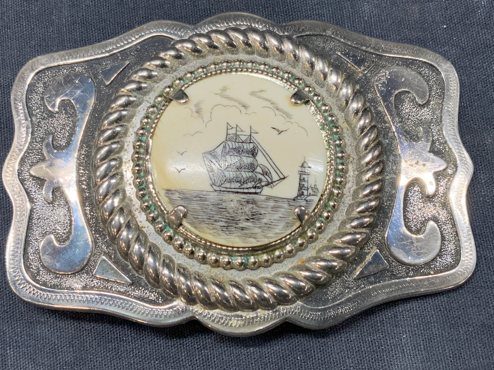 Scrimshaw Style Belt Buckle: Silver toned metal belt buckle with an etched nautical ship center piece, possibly made of resin. Approx 4 x 3 inches and center design is approx 1.5 x 1.5 inches Moderate wearing to metal surface Scr