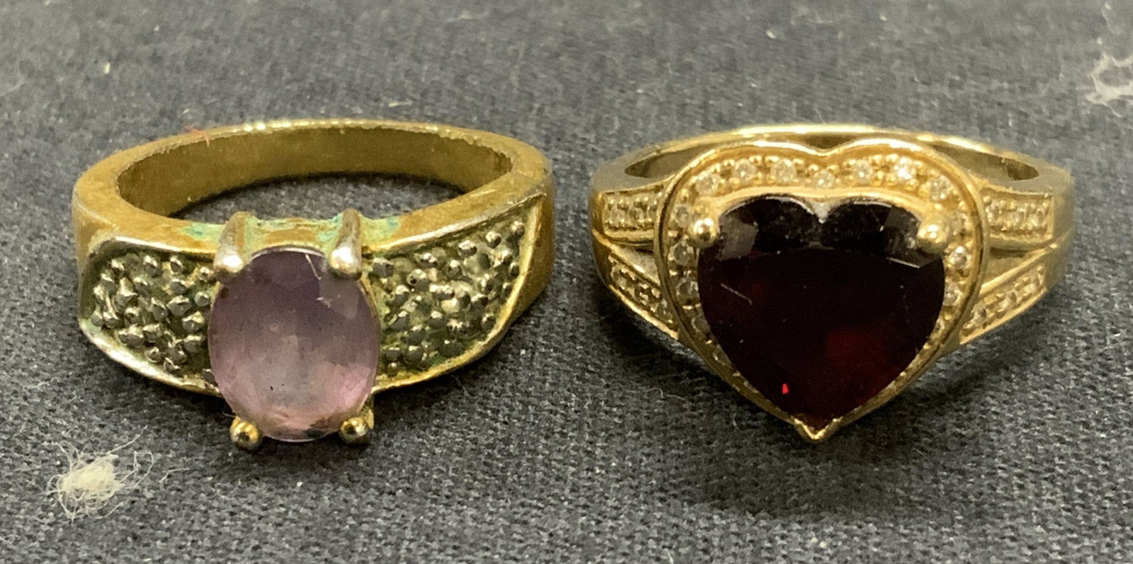 Lot 2 Gold Tone Amethyst & Garnet Rings: Lot of 2 jewelry pieces. Gold tone ring with amethyst and colorless gemstones. Ring size 8. Gold tone ring with garnet and colorless gemstones. Ring size 8.5. Both pieces have surface wear including s