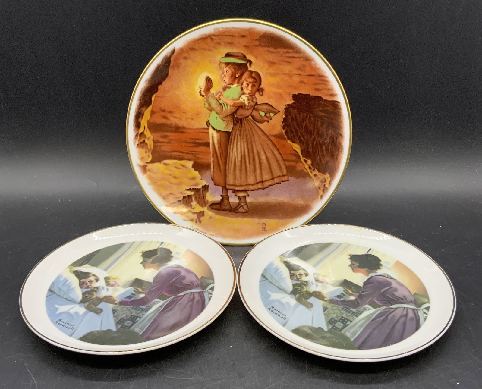 Lot 3 Vintage Norman Rockwell Porcelain Plates: Three vintage porcelain plates. Imagery to plates primarily depicts children. Two are the same and feature a mother looking after her two children. Largest measures approx. 7.75L x 7.75W in. Underside